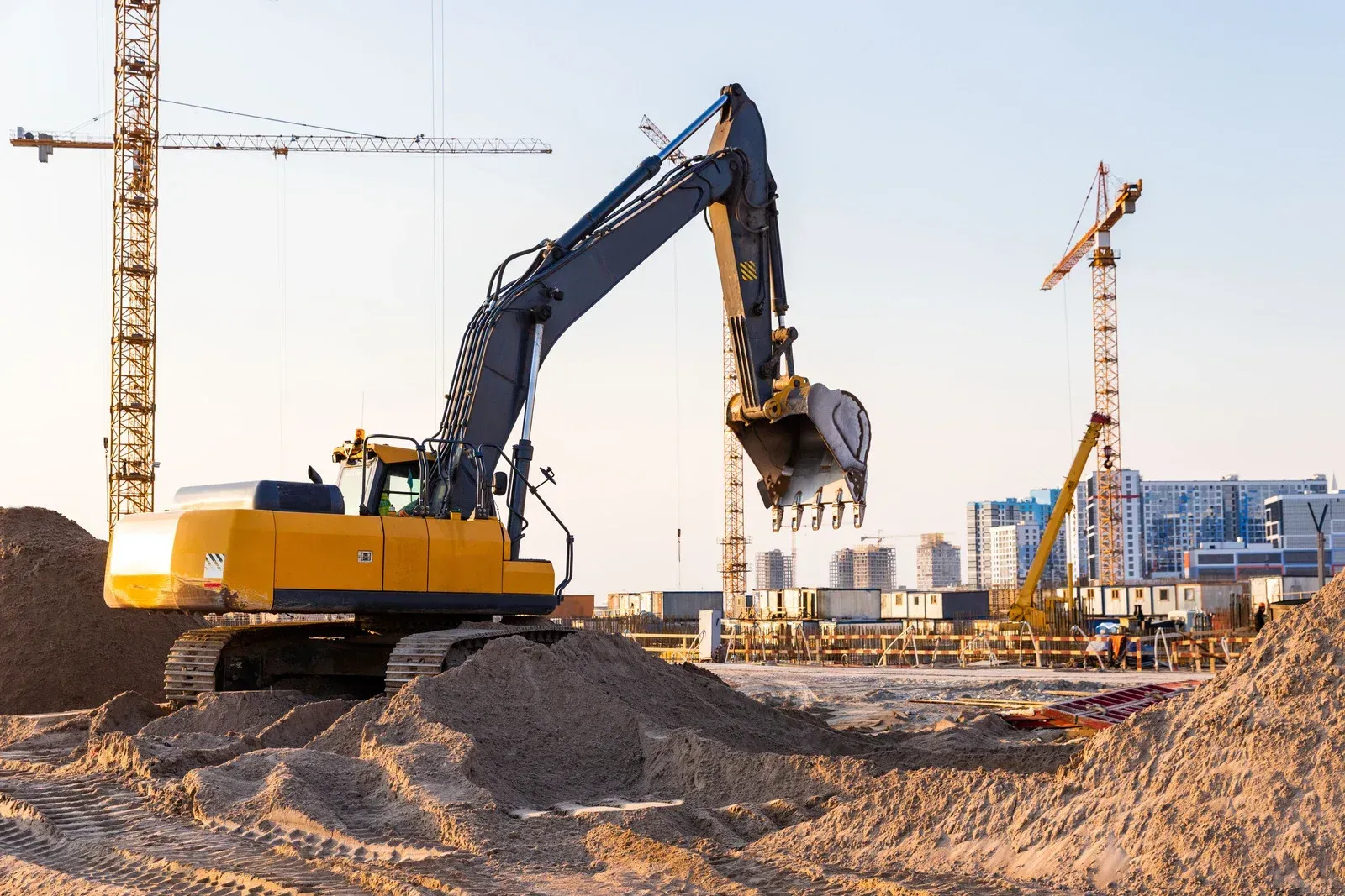 Yellow excavator at a construction site with cranes and building materials in the background