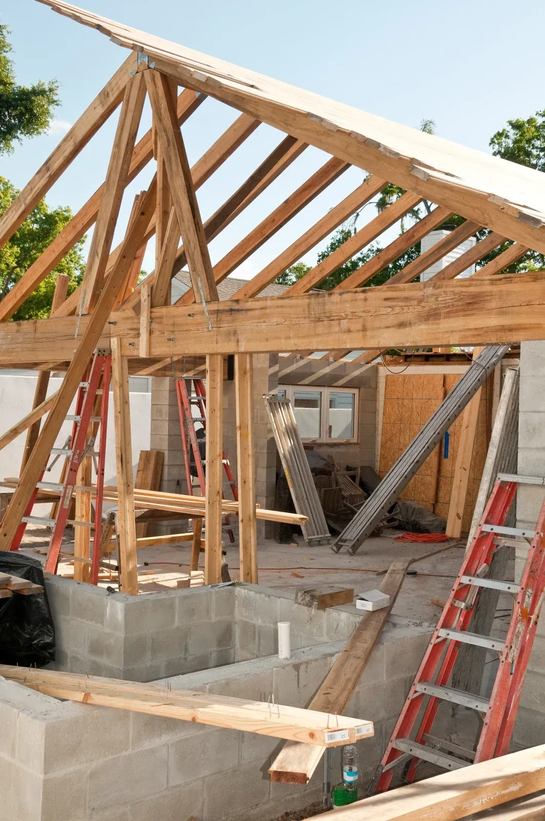 Wood-framed house under construction with roof trusses, ladders, and cinder block foundation