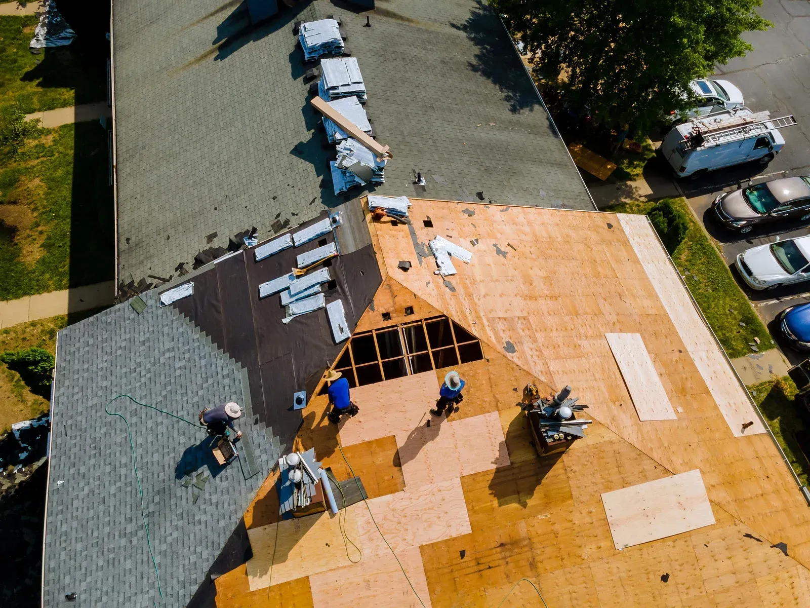 Workers installing roof shingles on a house, with exposed plywood and gray and tan sections.