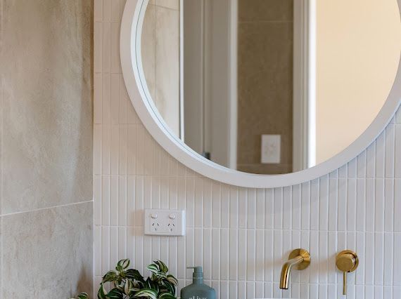 Bathroom with round white mirror, gold fixtures, and tiled wall. — Flow Kitchens in Taree South, NSW