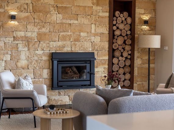 Fireplace in a stone wall, flanked by wall sconces. Log storage niche, seating, and a side table are also present. — Flow Kitchens in Taree South, NSW