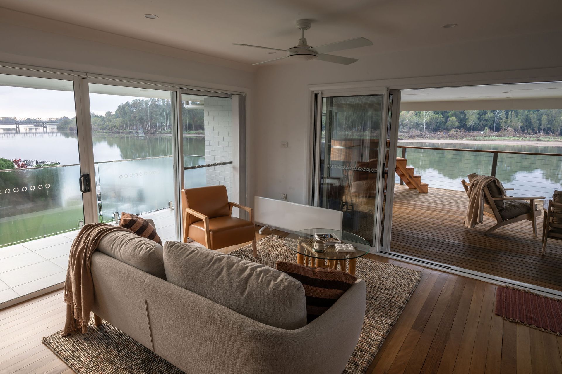 Living room with lake view, large sliding doors open to deck with chairs, sofa, brown and white tones. — Flow Kitchens in Taree South, NSW