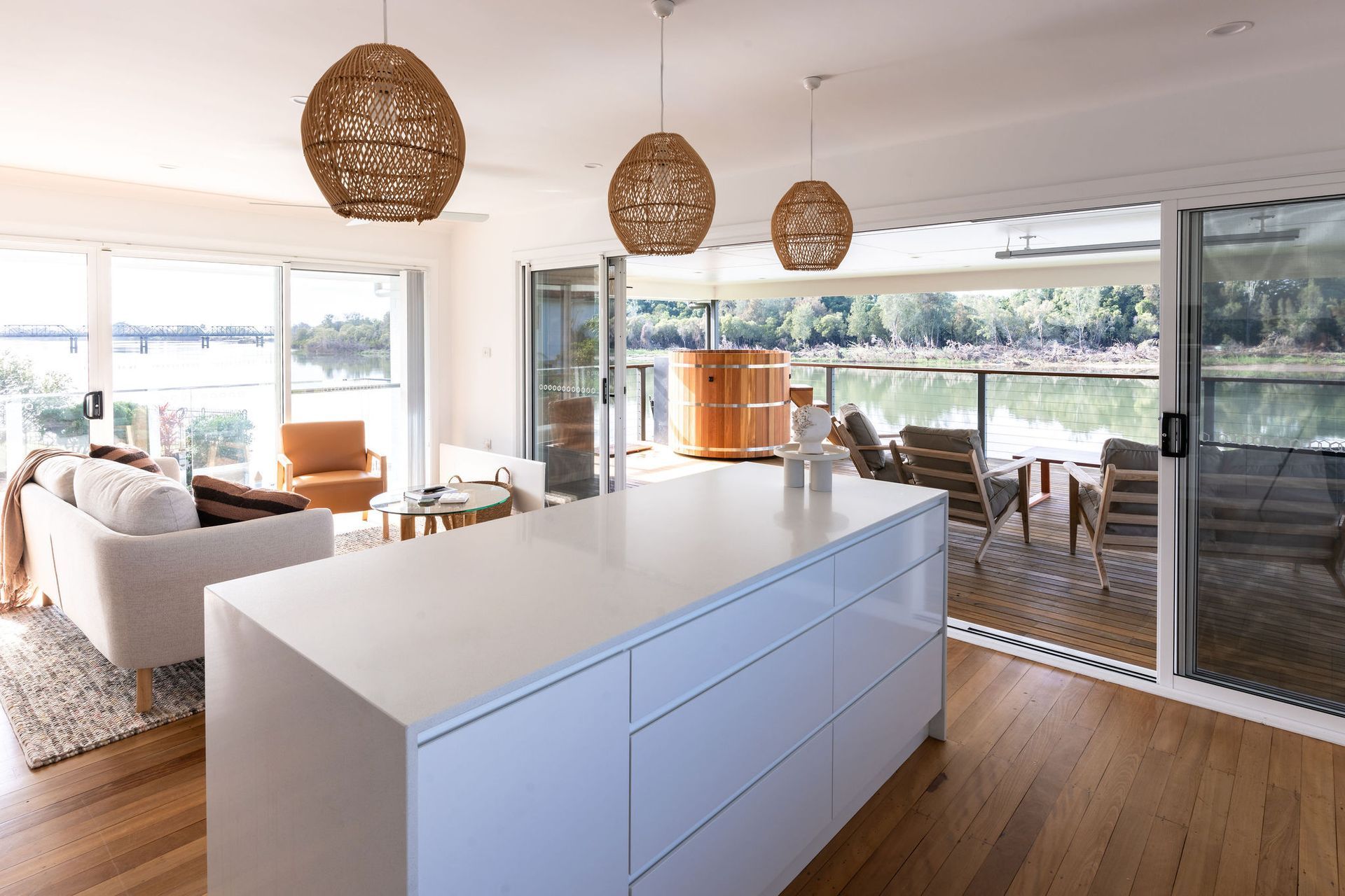 Bright kitchen with white island, woven pendant lights, and open to a lakeside deck. — Flow Kitchens in Taree South, NSW