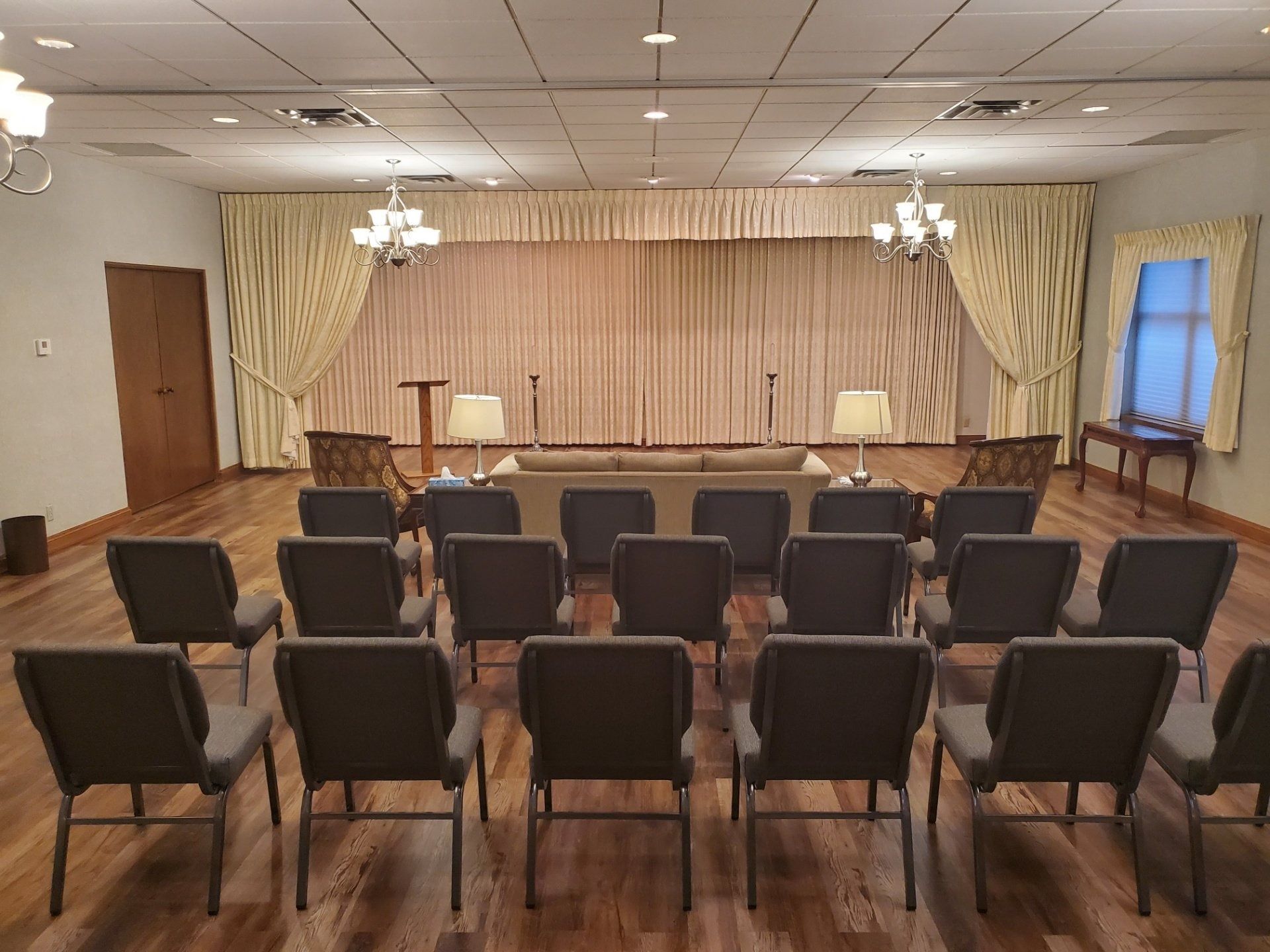 A funeral home viewing room with rows of chairs facing a stage with a curtain and sofa.