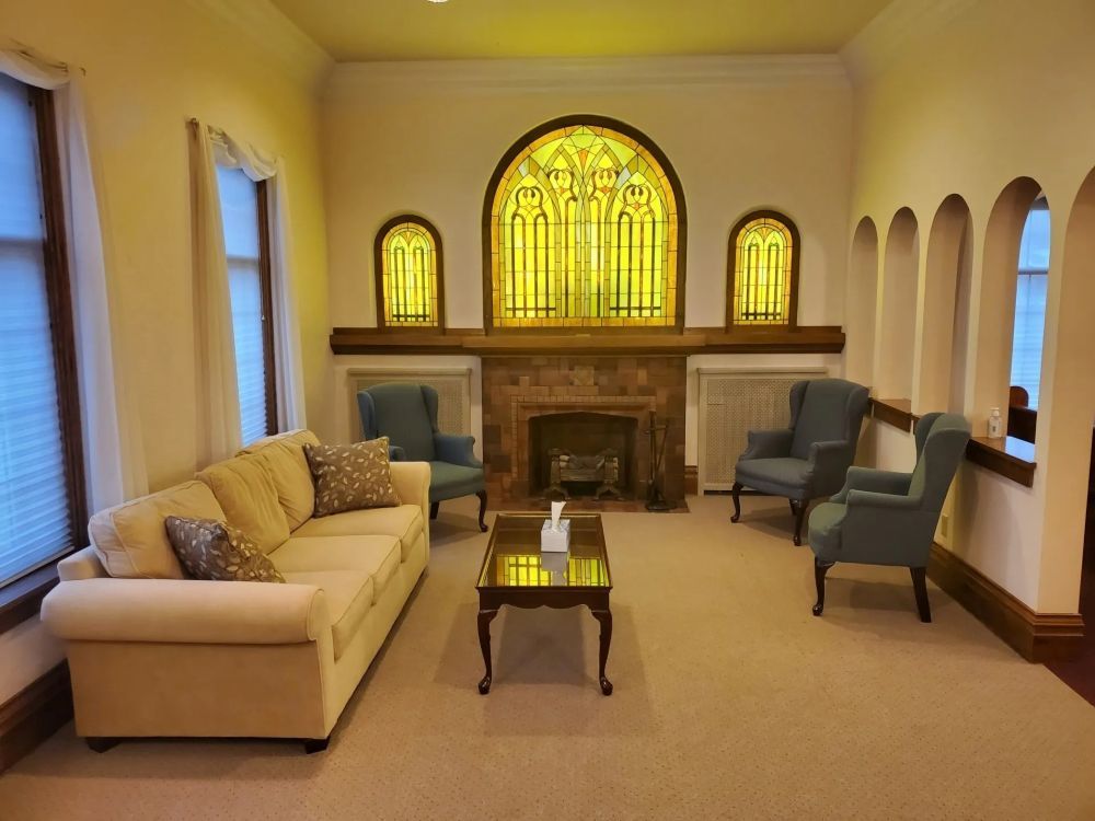 Cozy room with stained glass above a fireplace, flanked by arched alcoves. Beige sofa, chairs, and rug.
