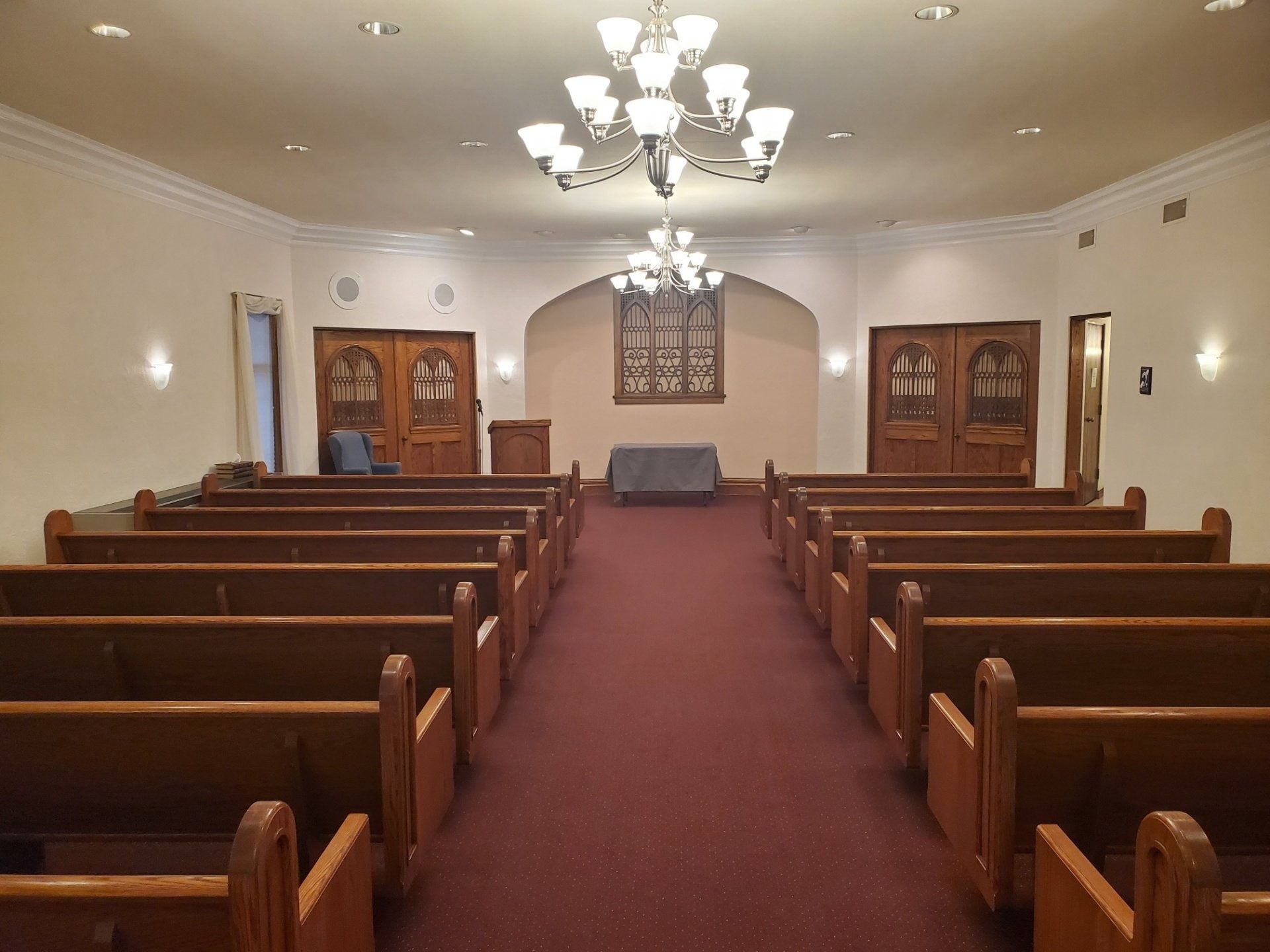 Interior of a chapel with wooden pews, red carpet, and two chandeliers.