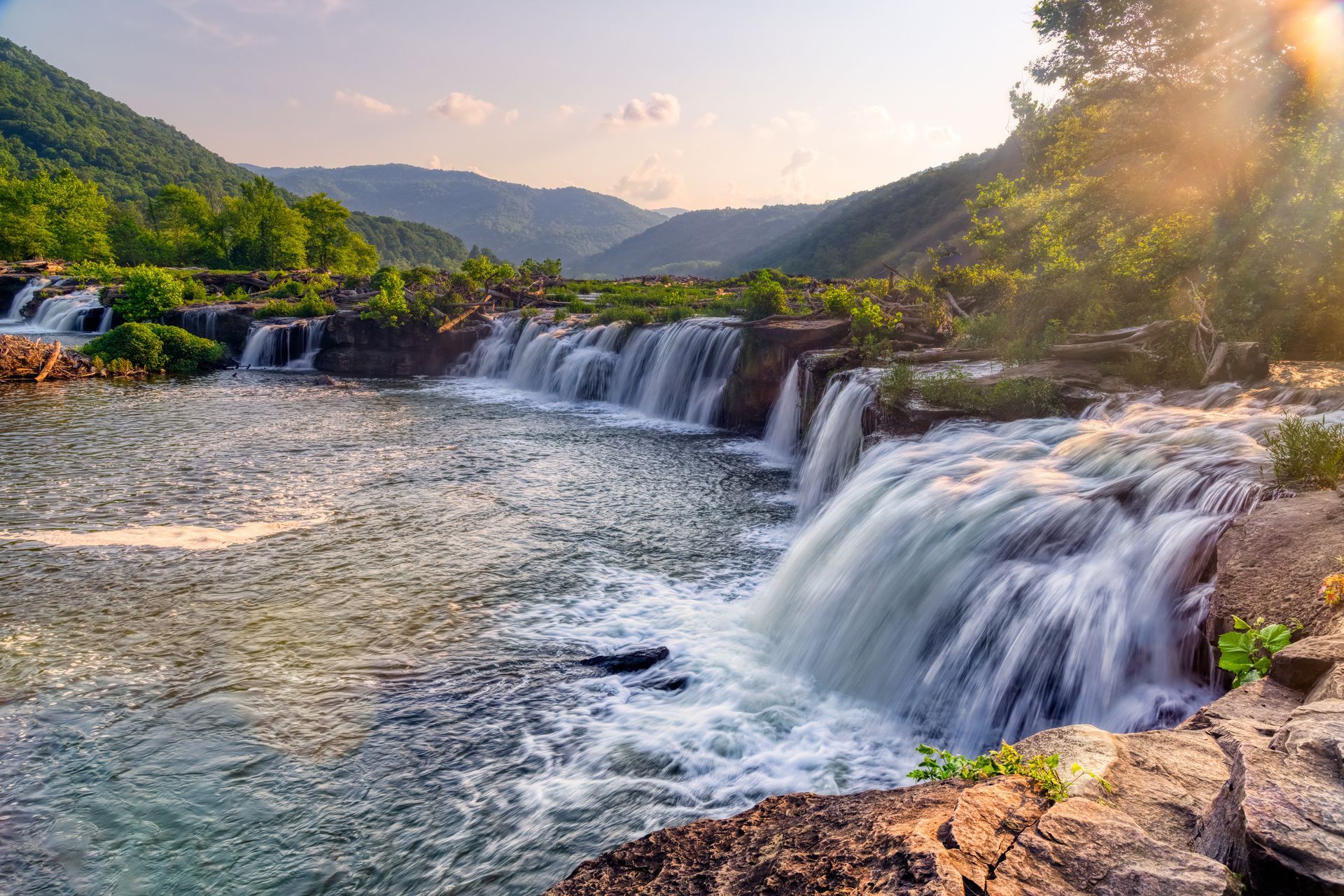 A waterfall in the middle of a river with mountains in the background.