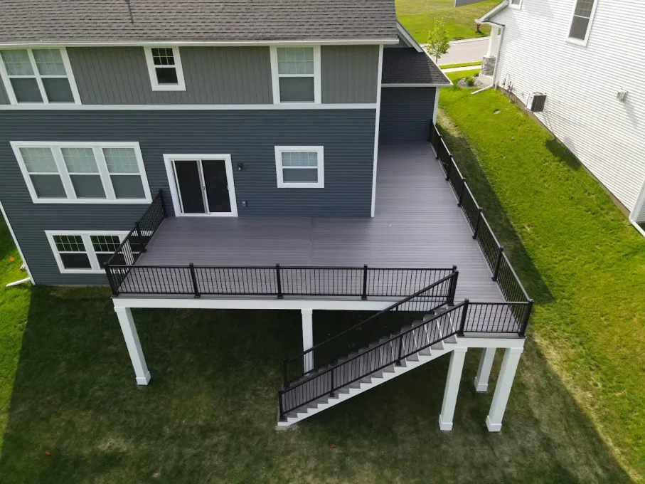 A high-angle view of a grey composite deck with black railings and stairs attached to the back of a blue-sided house.