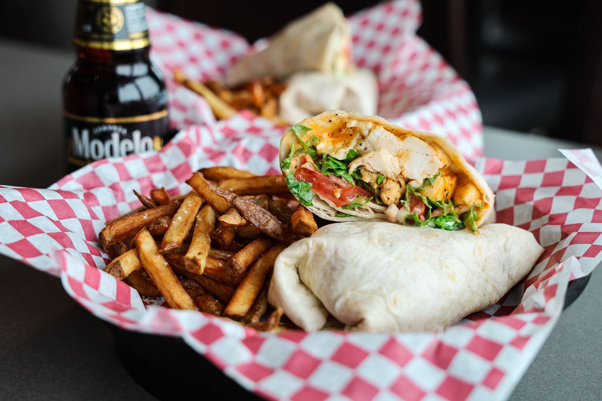 A sandwich and french fries in a basket with a bottle of modelo beer in the background.