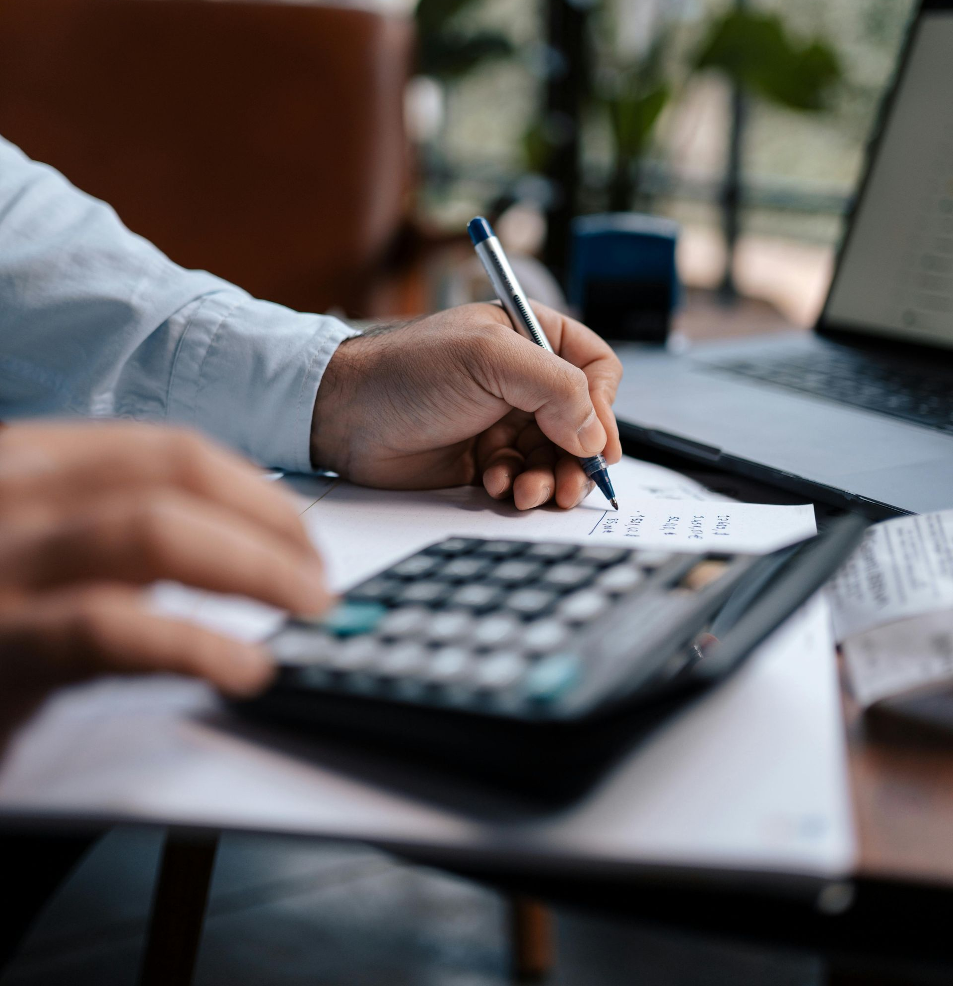 Person using a calculator and pen to write on paper near a laptop.