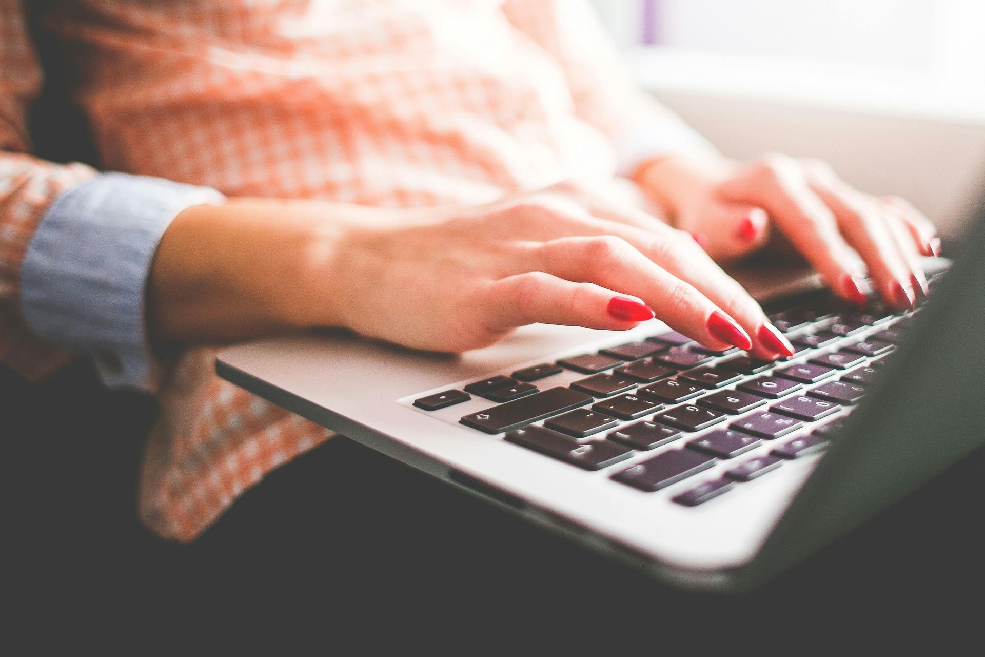 Woman typing on a laptop; red nail polish, wearing a pink patterned shirt.
