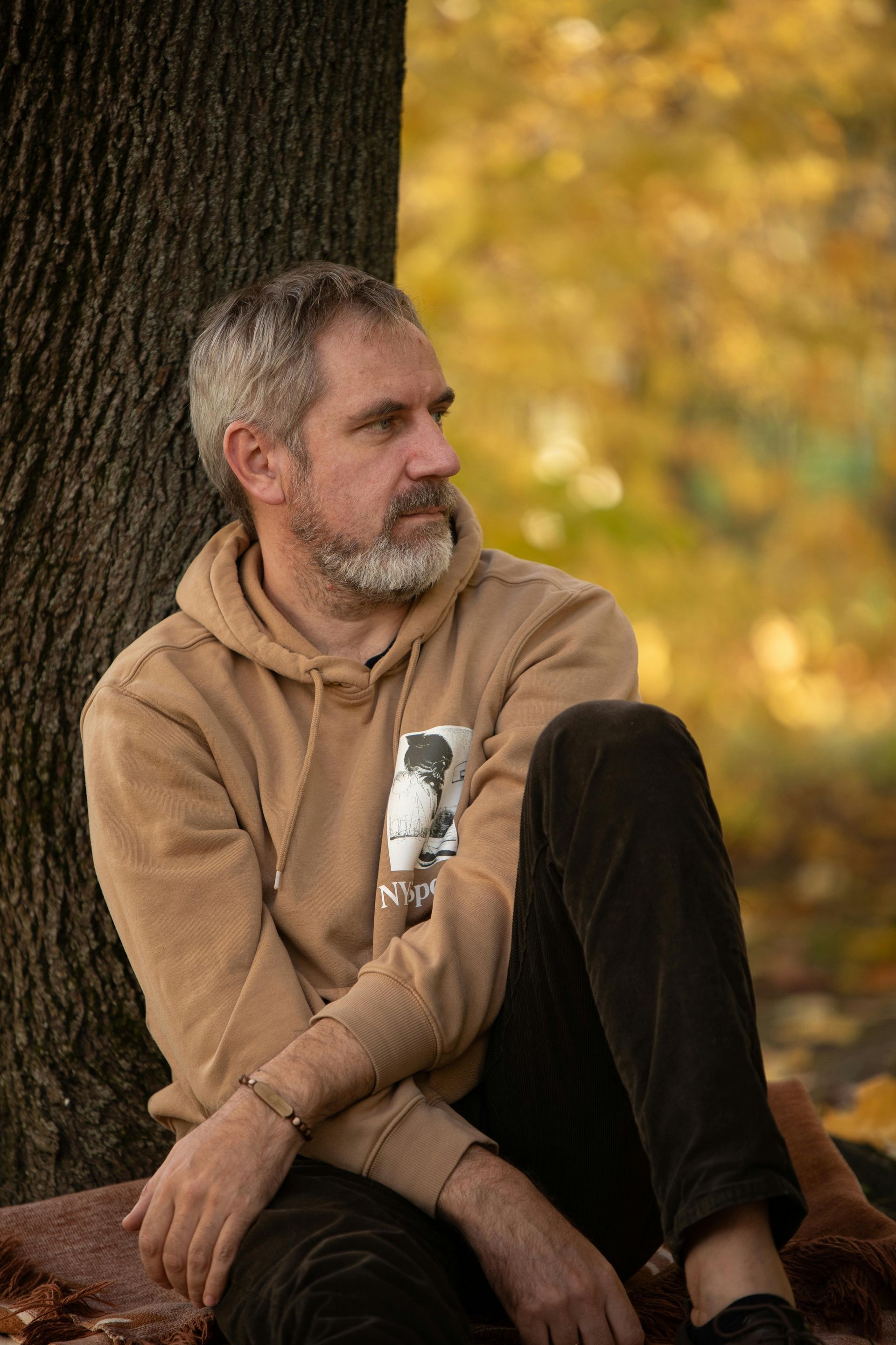 Man in tan hoodie sits by tree, looking away; fall setting with yellow leaves.