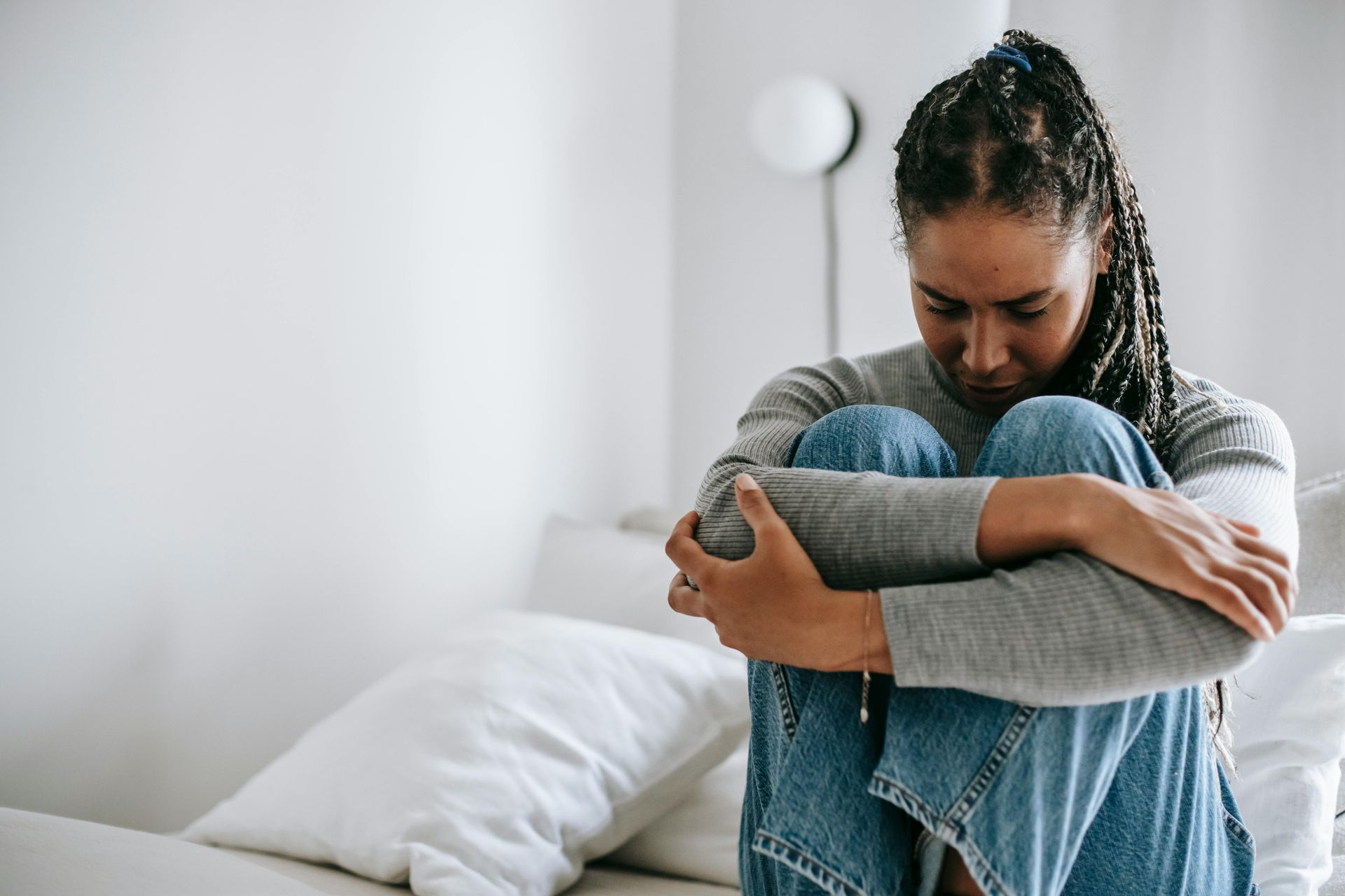Woman in jeans and grey sweater sits on a bed, hugging her knees, looking down with a sad expression.