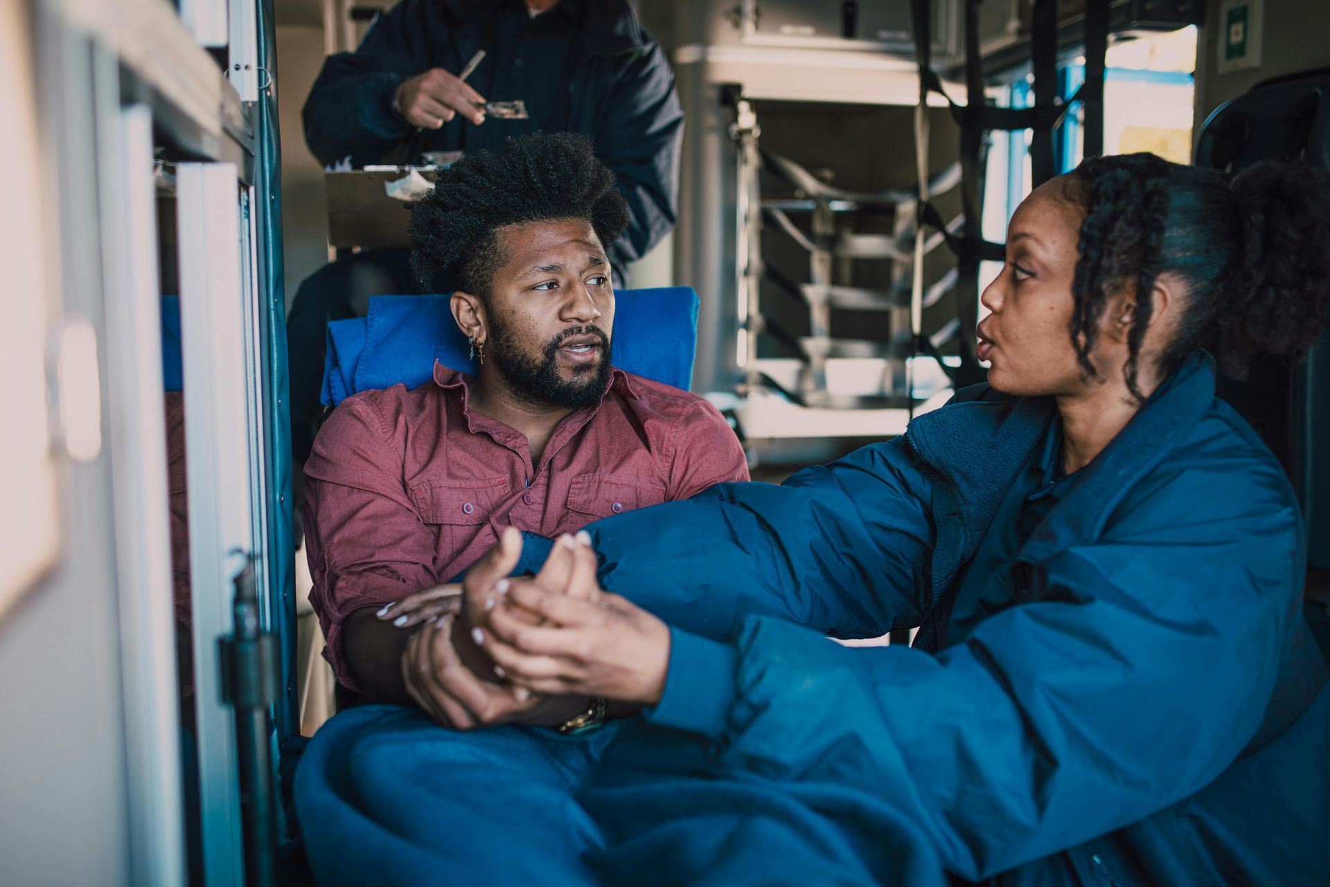 Man and woman in ambulance, talking. Man looks concerned, woman holds his hands. Third person works in background.