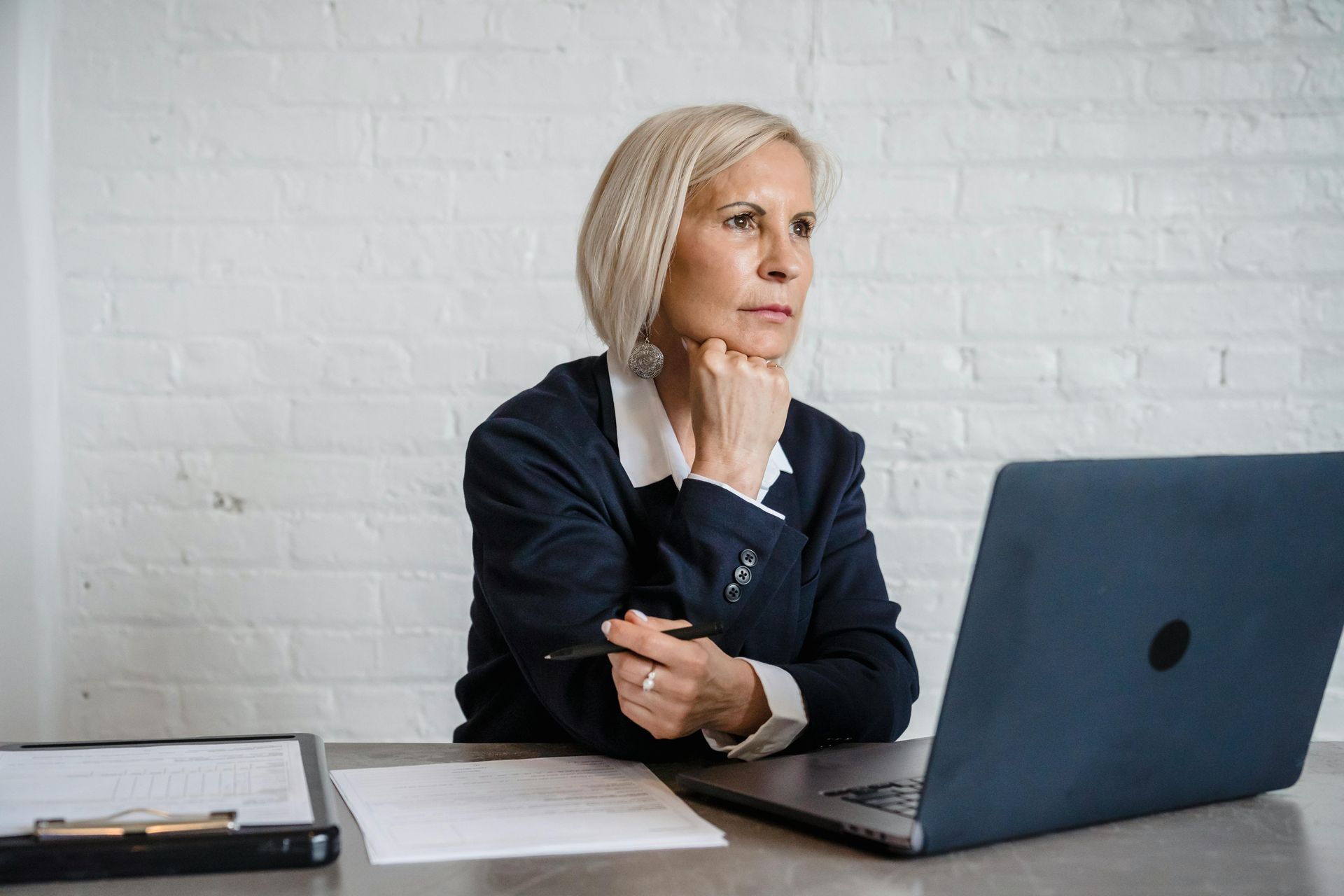 Woman with blonde hair, in a blazer, looking thoughtful at a laptop in a white-walled office.