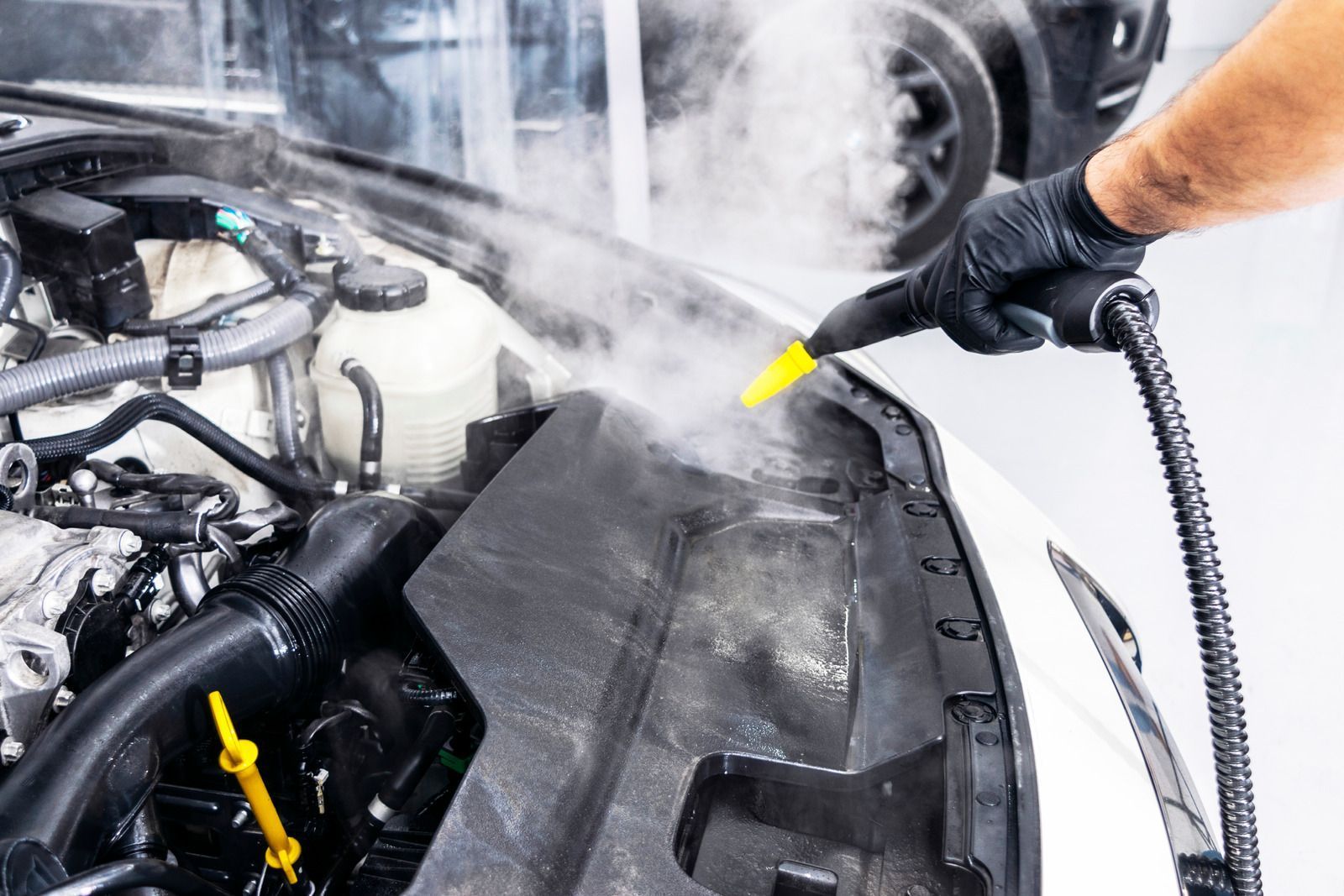 Steam cleaning a car engine. A gloved hand uses a yellow nozzle to direct steam.
