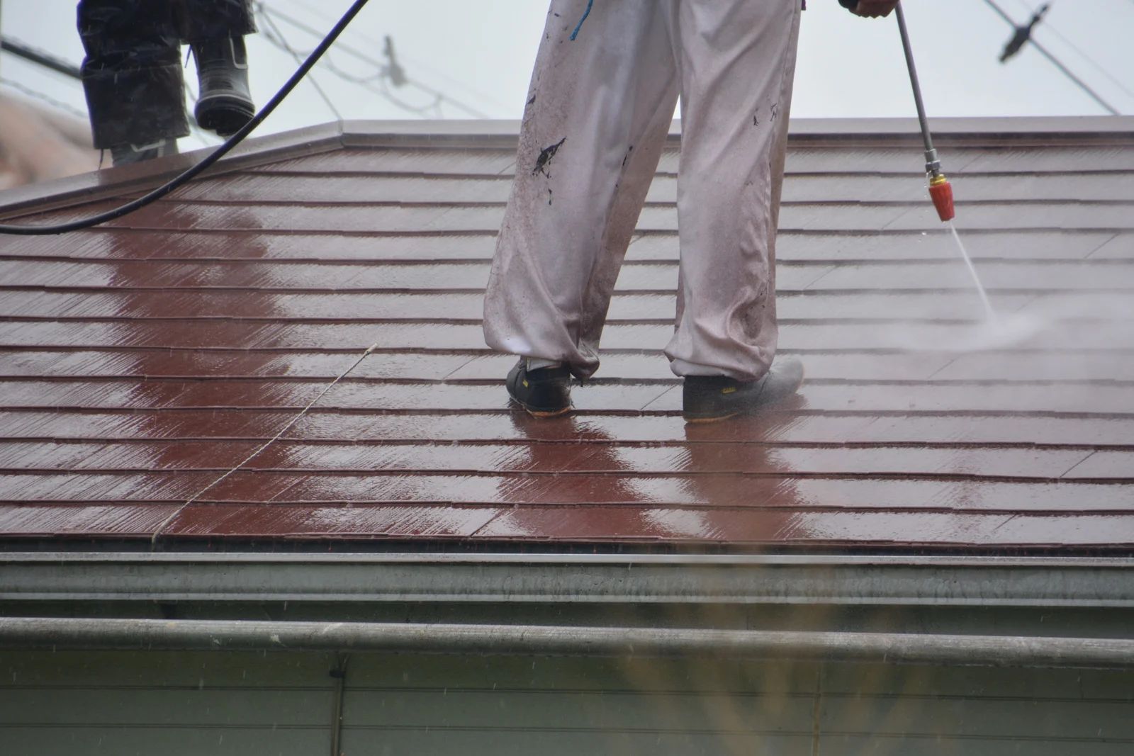 Two workers power washing a red tiled roof. One sprays water, the other stands nearby.