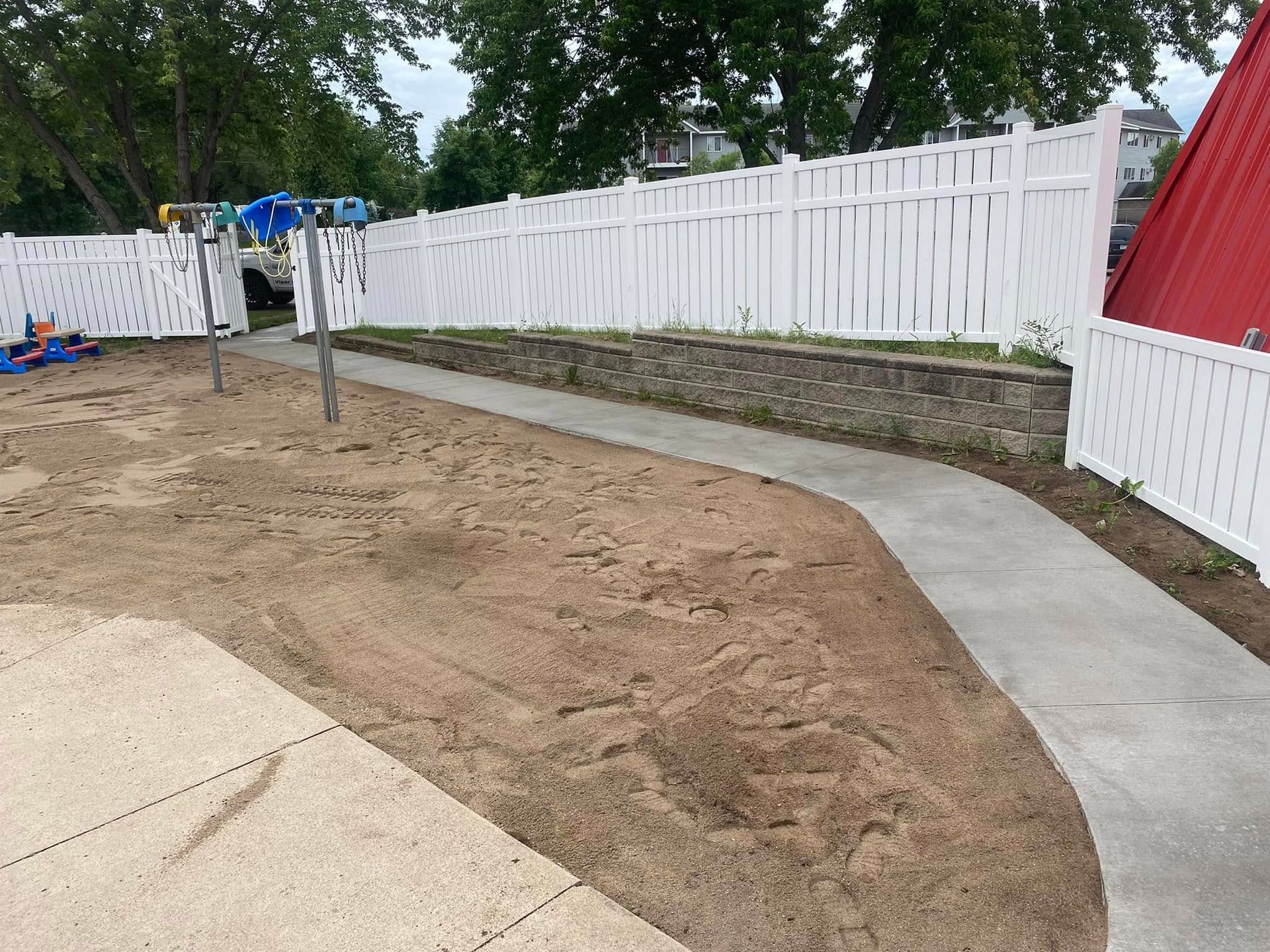 A concrete walkway leading to a playground with a white fence.