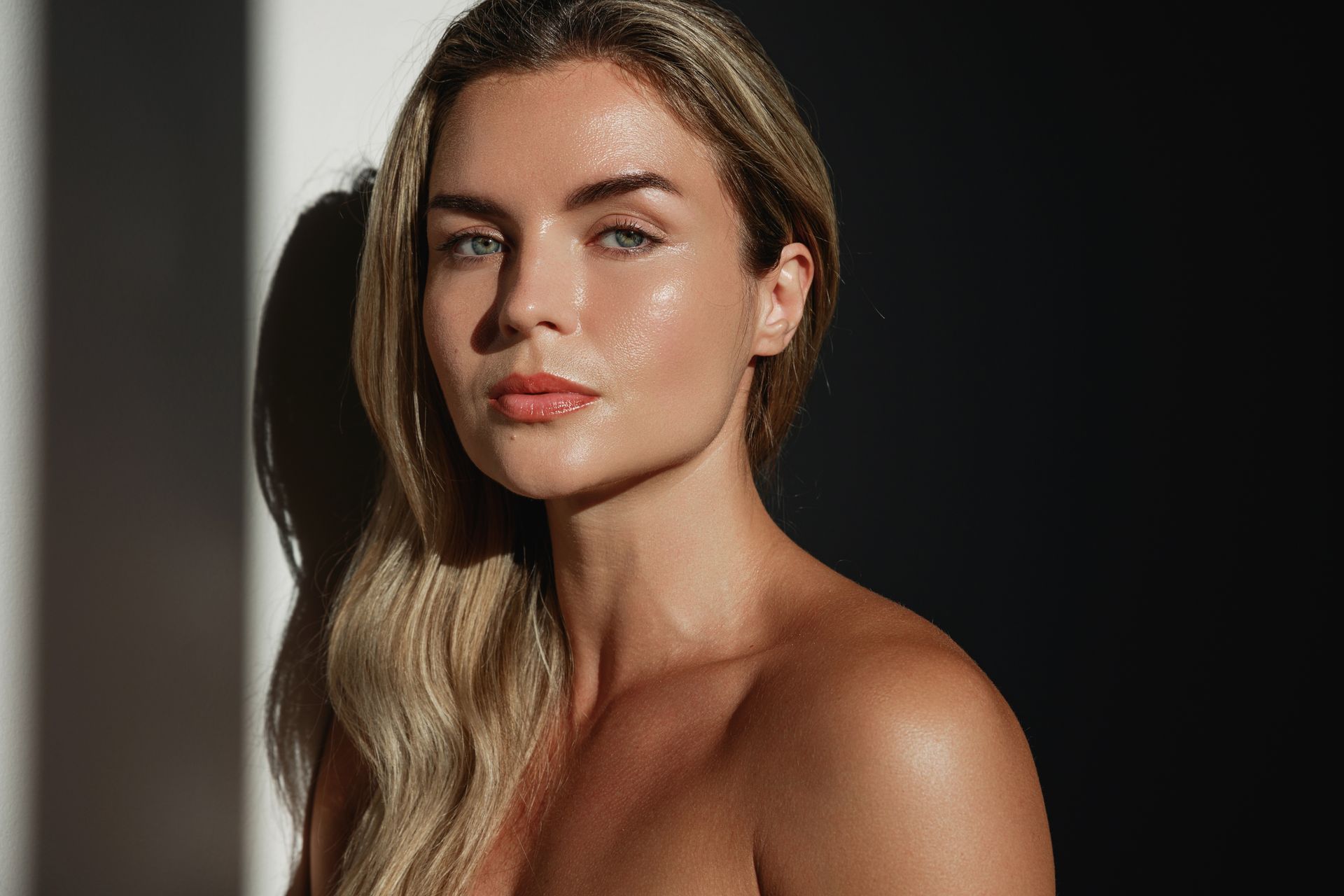 Woman with blonde hair, sun-kissed skin, in a dark setting with a shadow, looking at the camera.