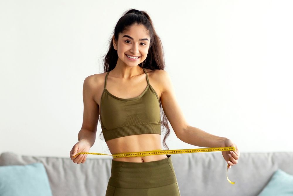 Woman in olive athletic wear smiles, measuring her waist with a yellow tape in a living room.