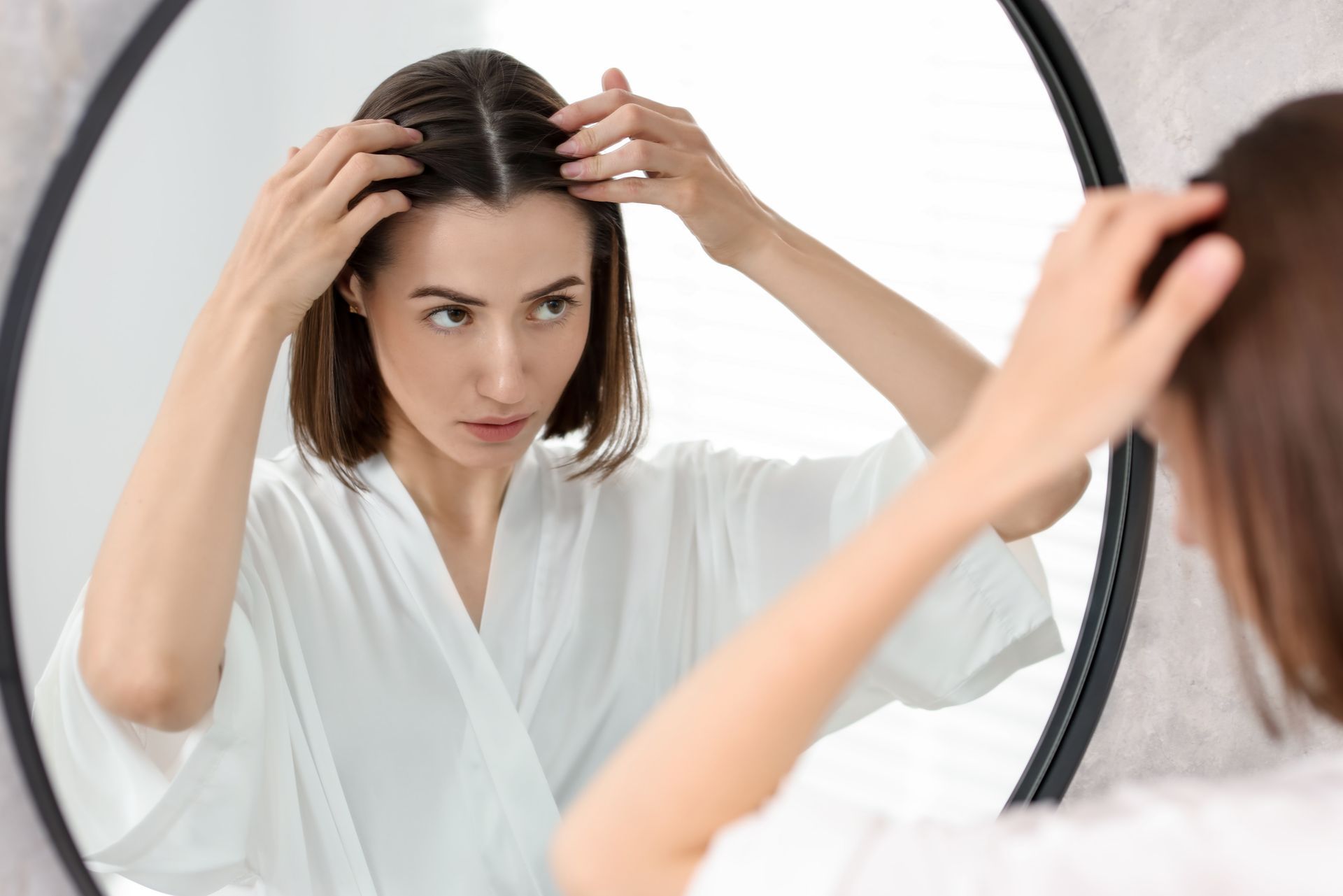 Woman in white robe examining her hair in a round mirror, indoors.