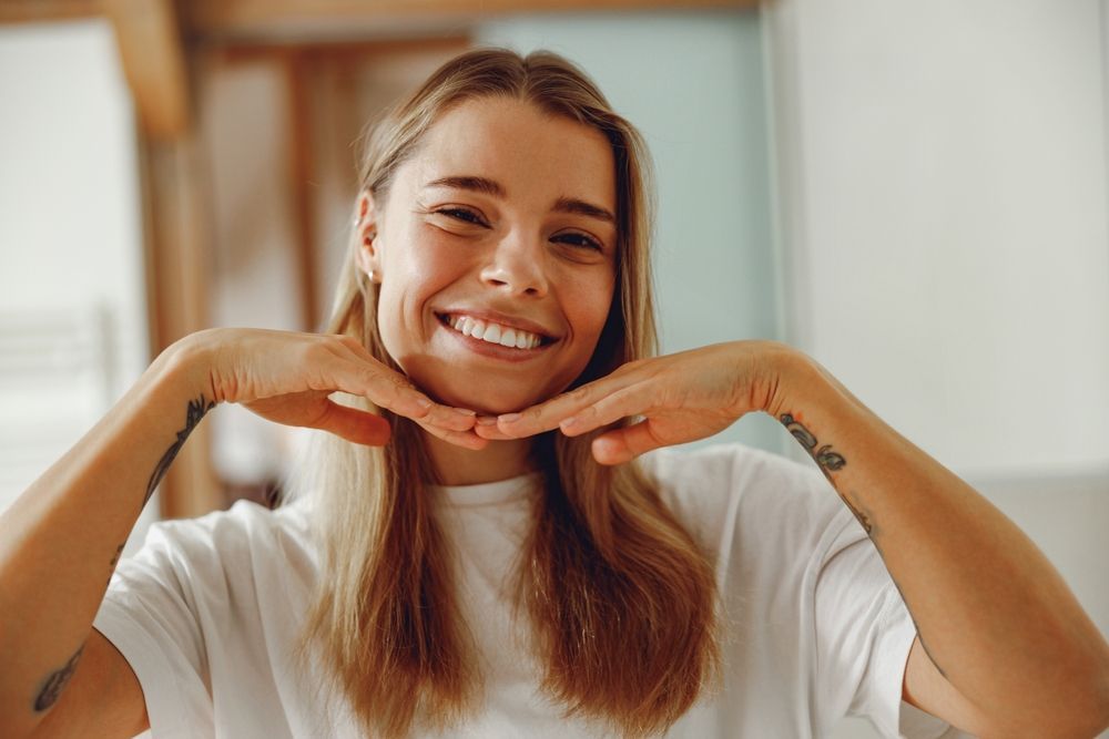 Blond person smiles, hands framing chin. Wearing white shirt, indoors.
