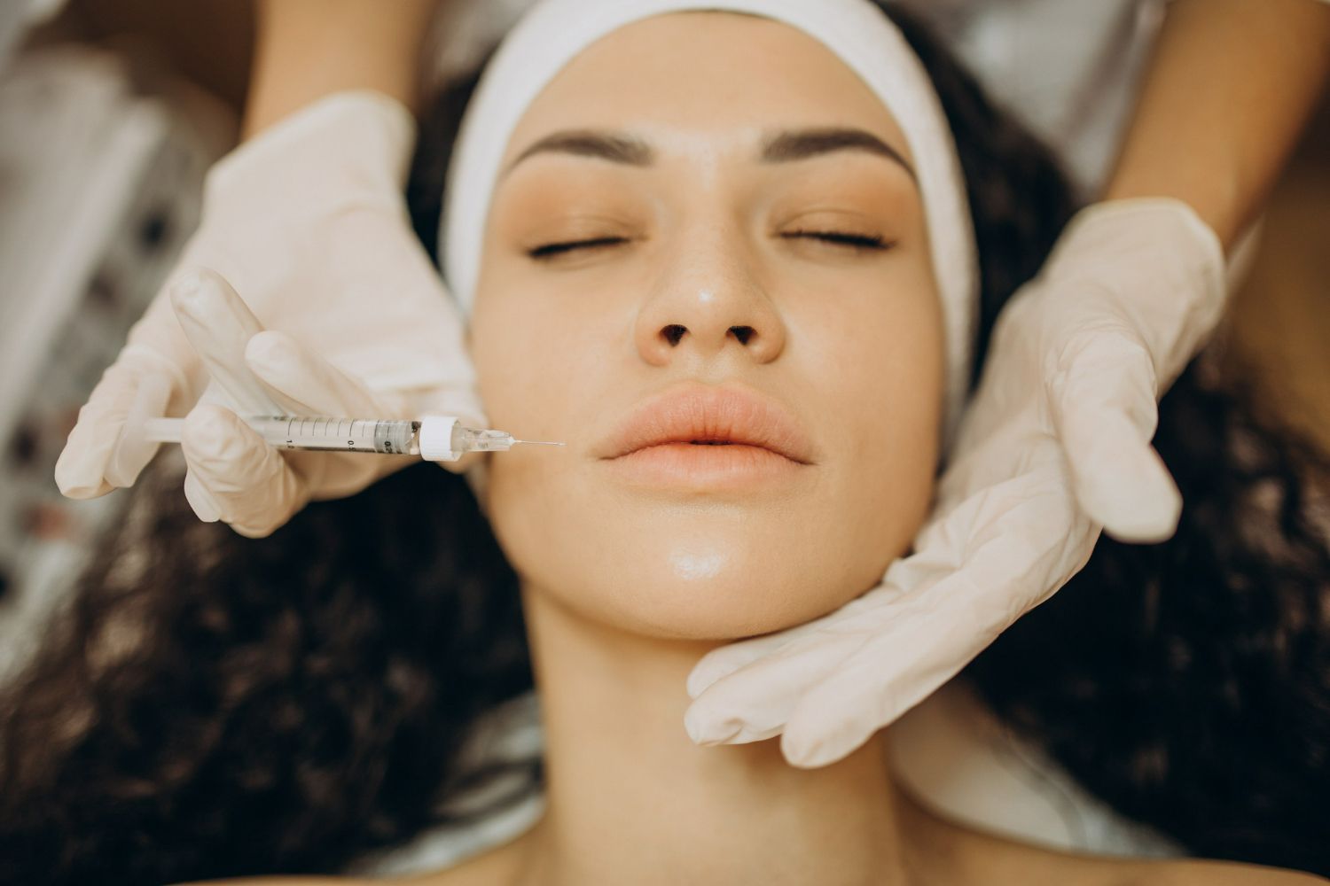 Woman receiving facial injections; gloved hands administering treatment near lips.