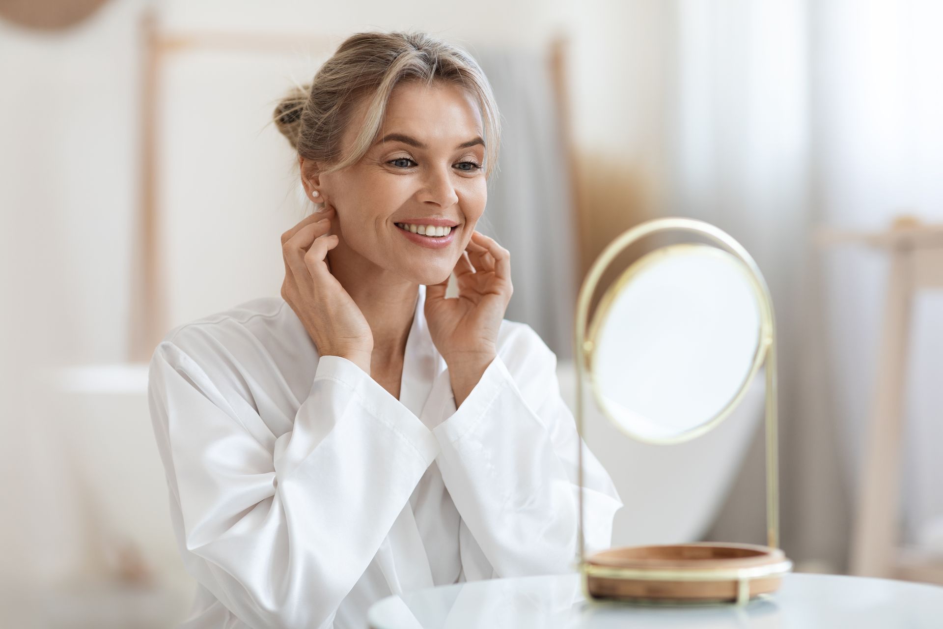 Woman in white robe looks in mirror, smiling, in bathroom.
