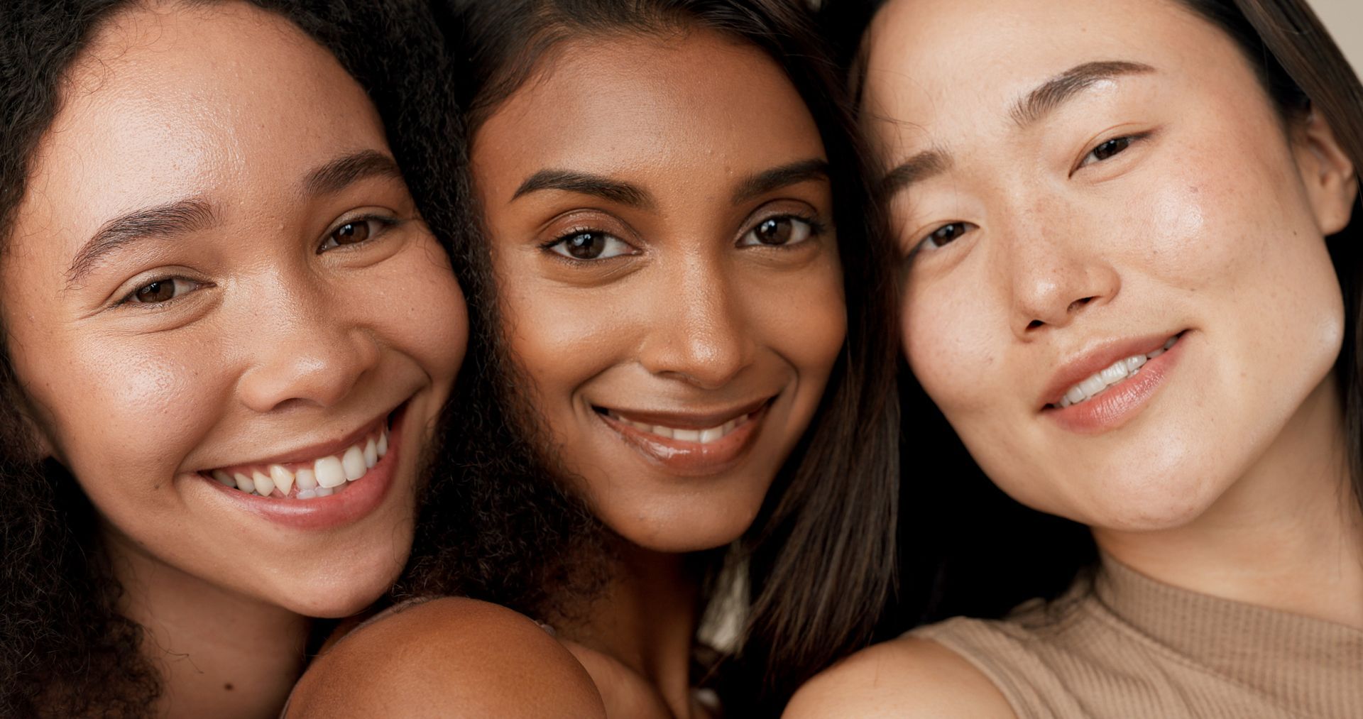 Three diverse women smiling close-up. Brown, tan, and Asian complexions are highlighted.