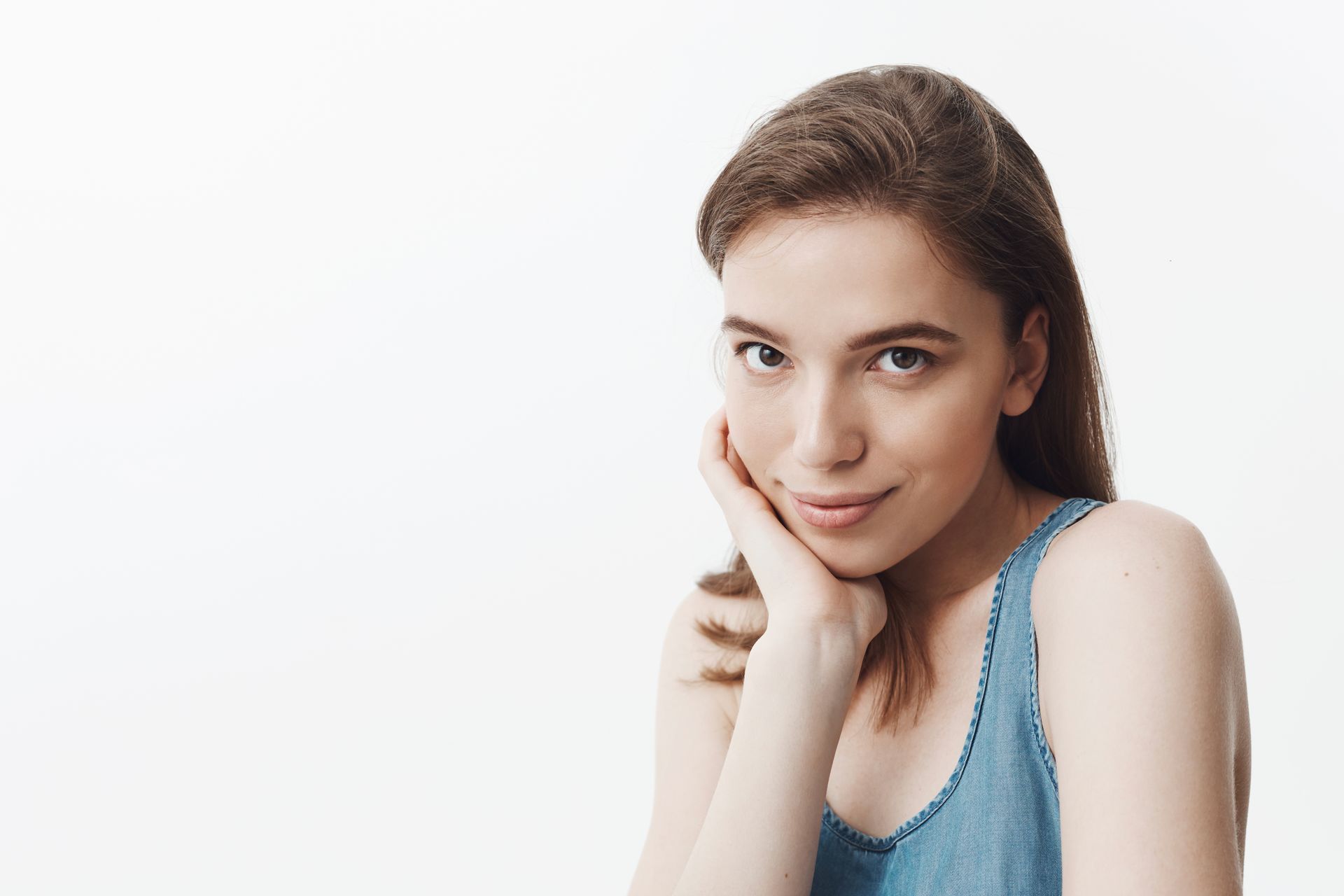 Woman in blue denim top, rests chin on hand, looking at the camera with a slight smile. White background.