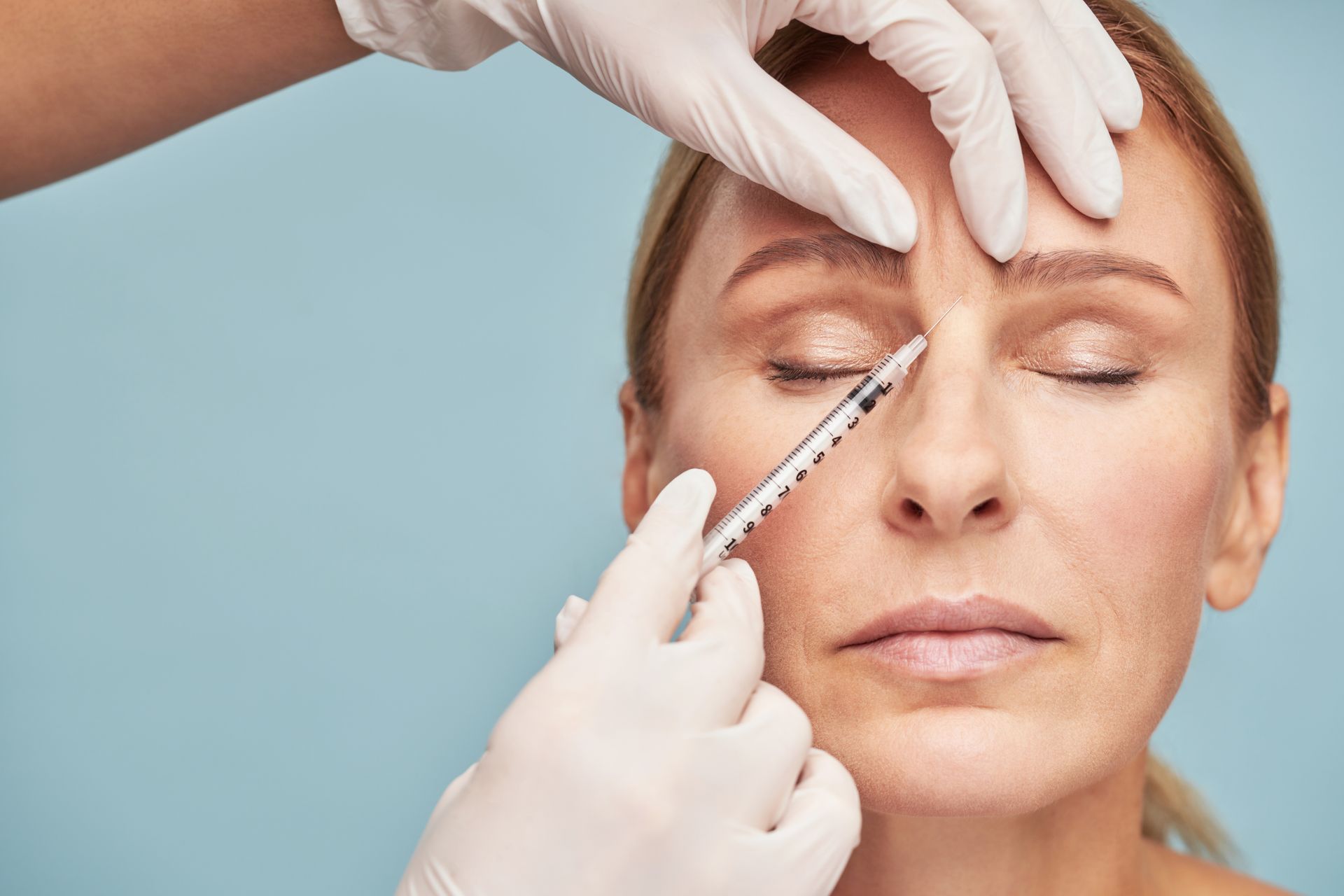 Gloved hands inject filler near a woman’s nose and brow during a cosmetic procedure