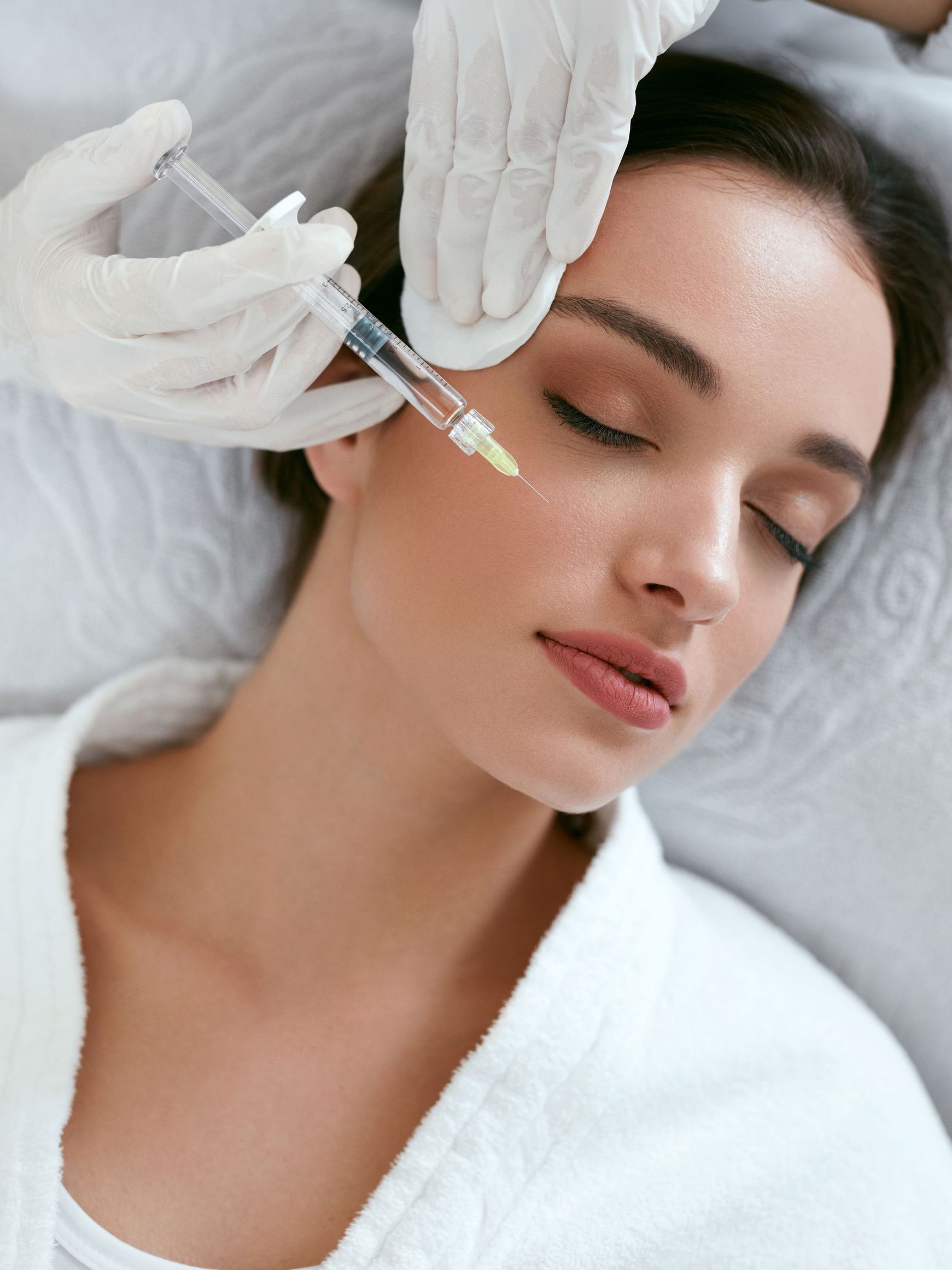 A woman receives a cosmetic injection near her eye. She is lying down with eyes closed, while a gloved hand injects her face.