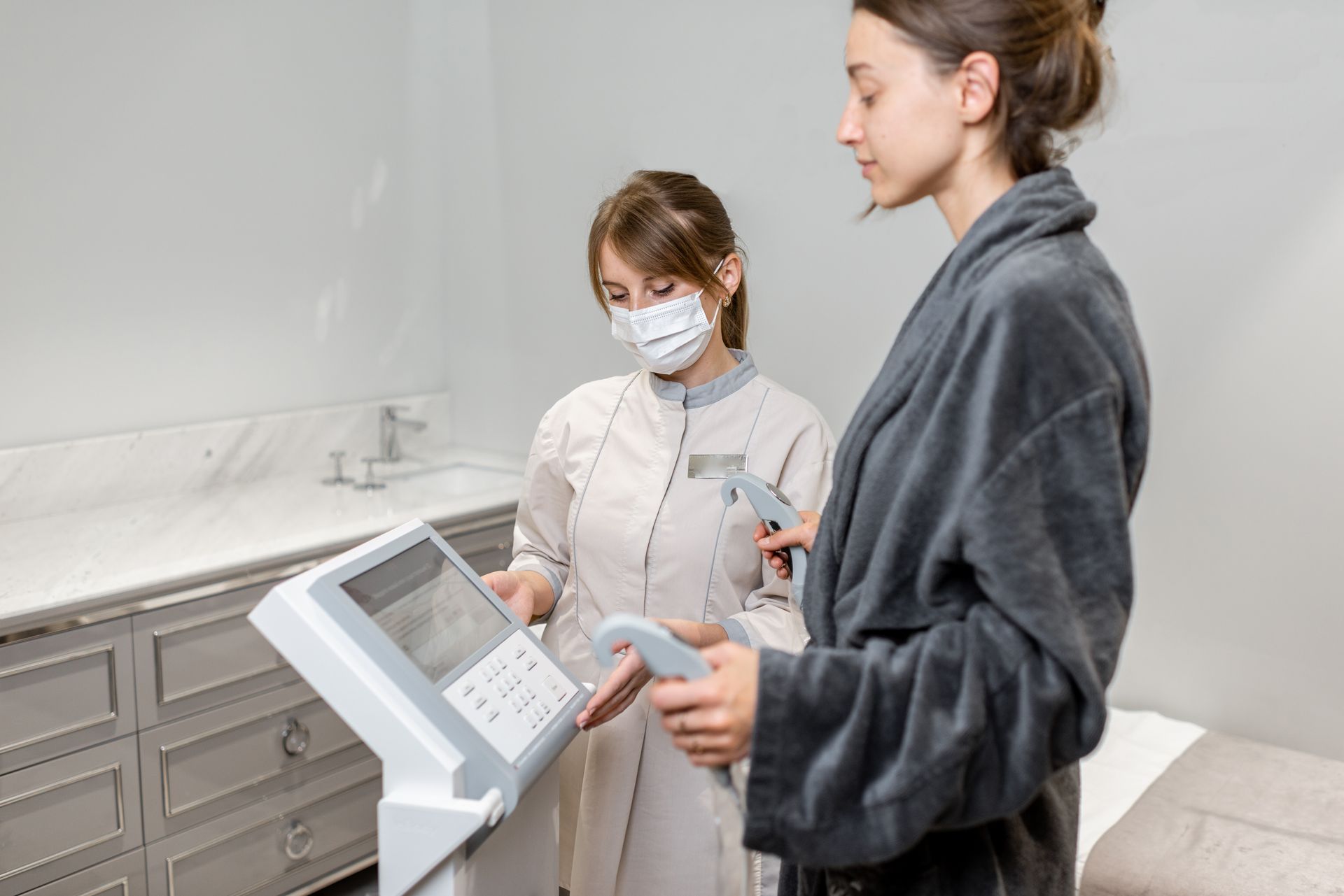 A woman in a robe stands next to a medical professional, who operates a body composition analyzer in a clinic.