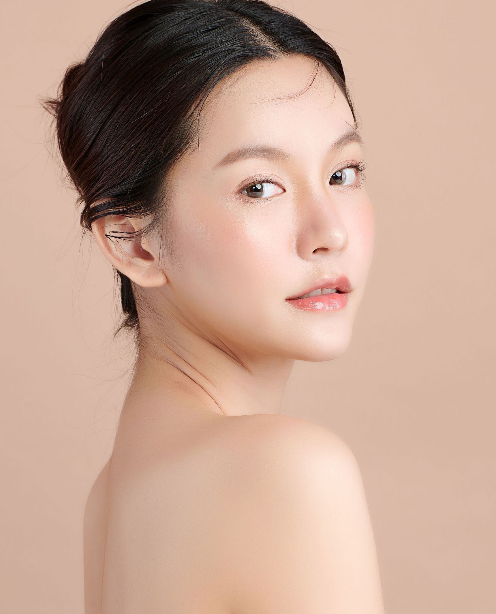 A light-skinned woman with slicked-back dark hair looks over her shoulder. She has a soft, glowing complexion against a peach background.