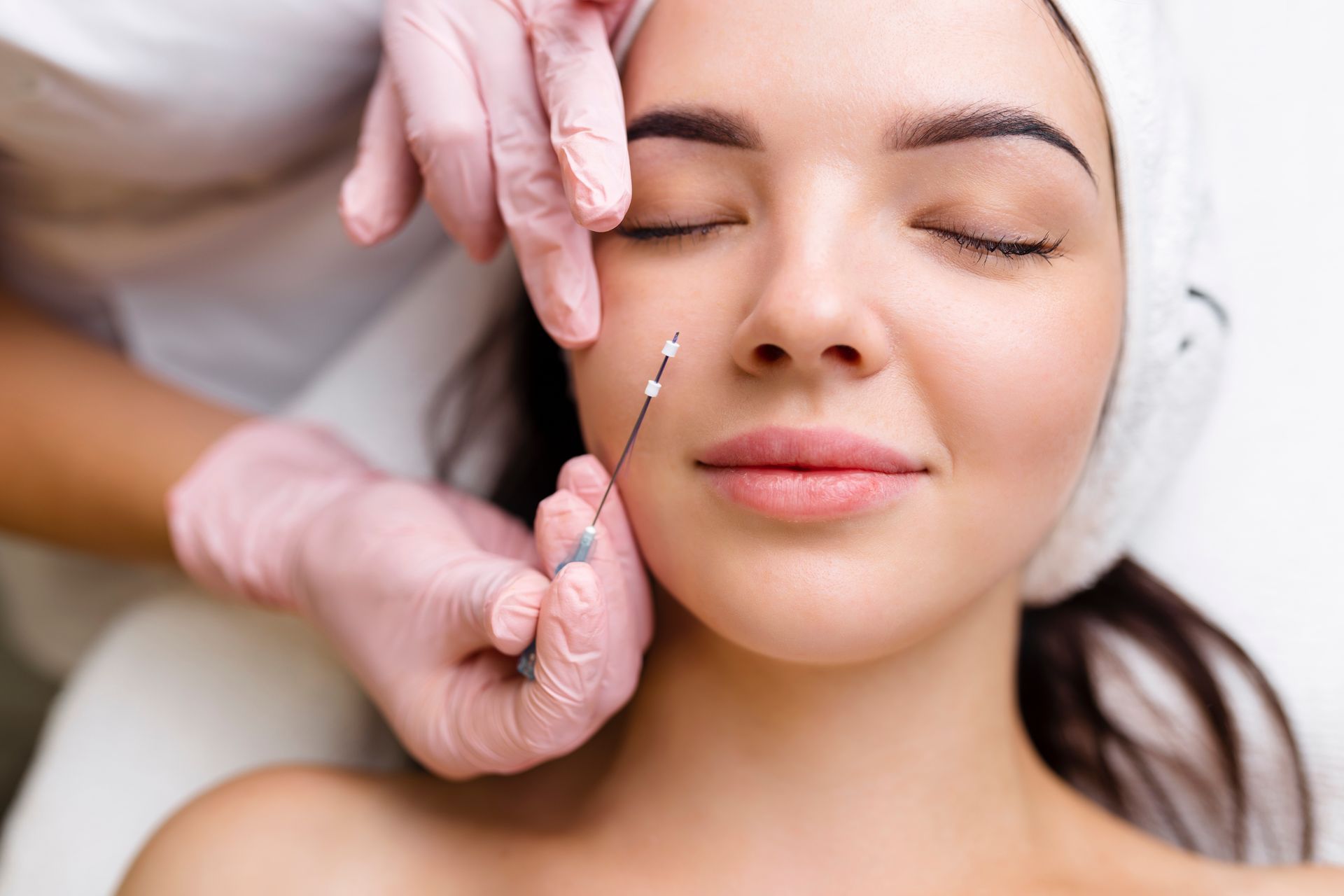 A person receiving a facial treatment, with a needle being held near their cheek. They have closed eyes.
