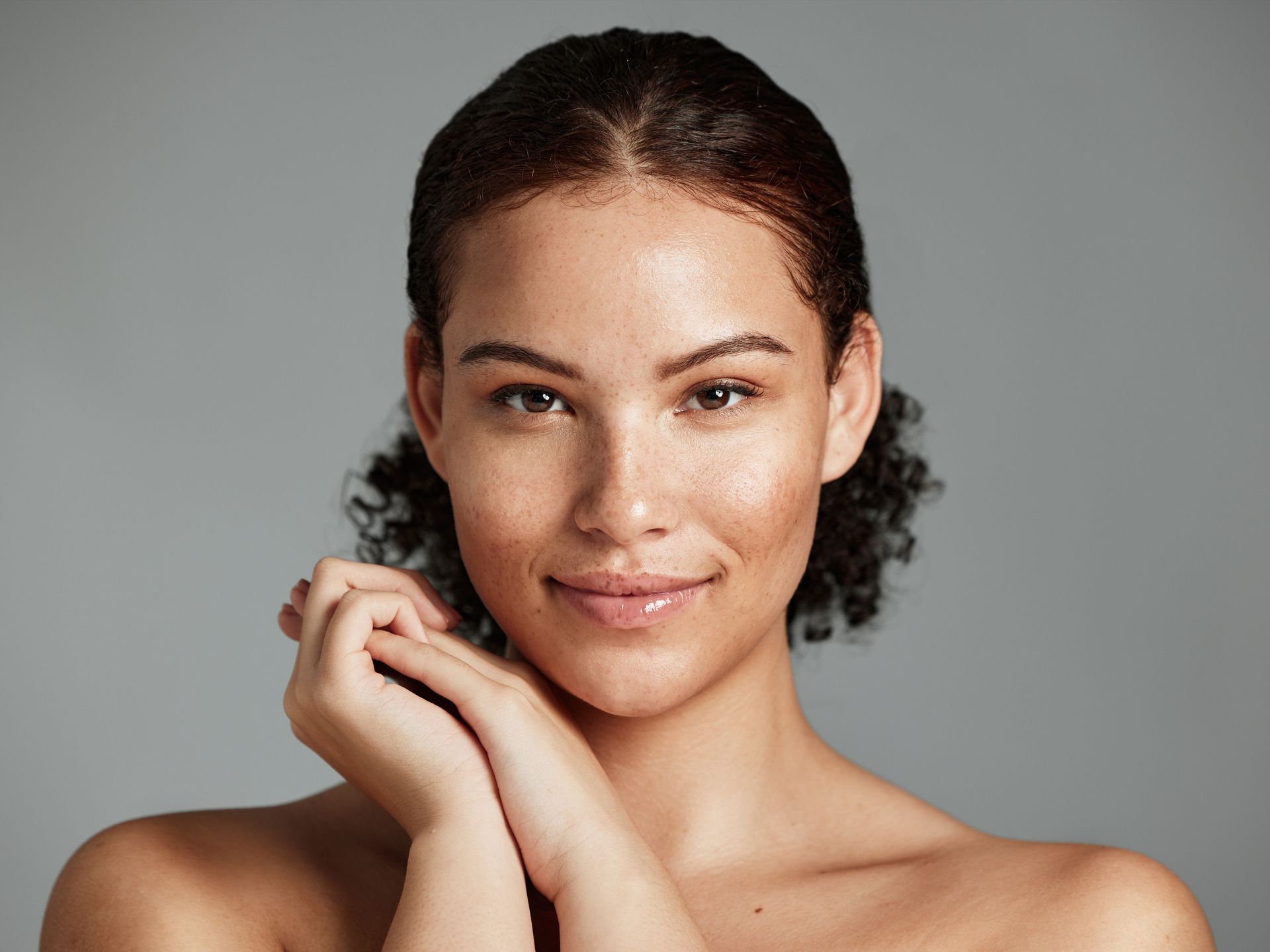 Smiling woman with curly dark hair, freckles, and hands clasped near her face, against a gray background.