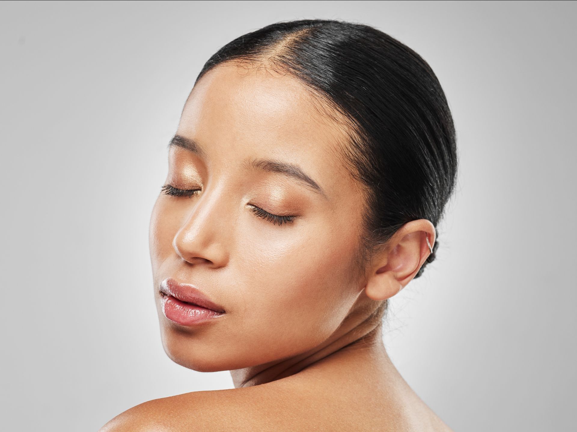 A woman with slicked-back dark hair and closed eyes. She has light brown skin and is looking over her shoulder, with a neutral background.