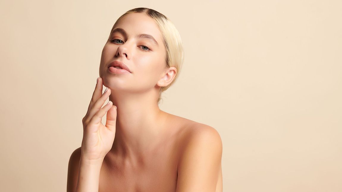 Blonde woman with bare shoulders, touching her neck and looking up, against a beige background.