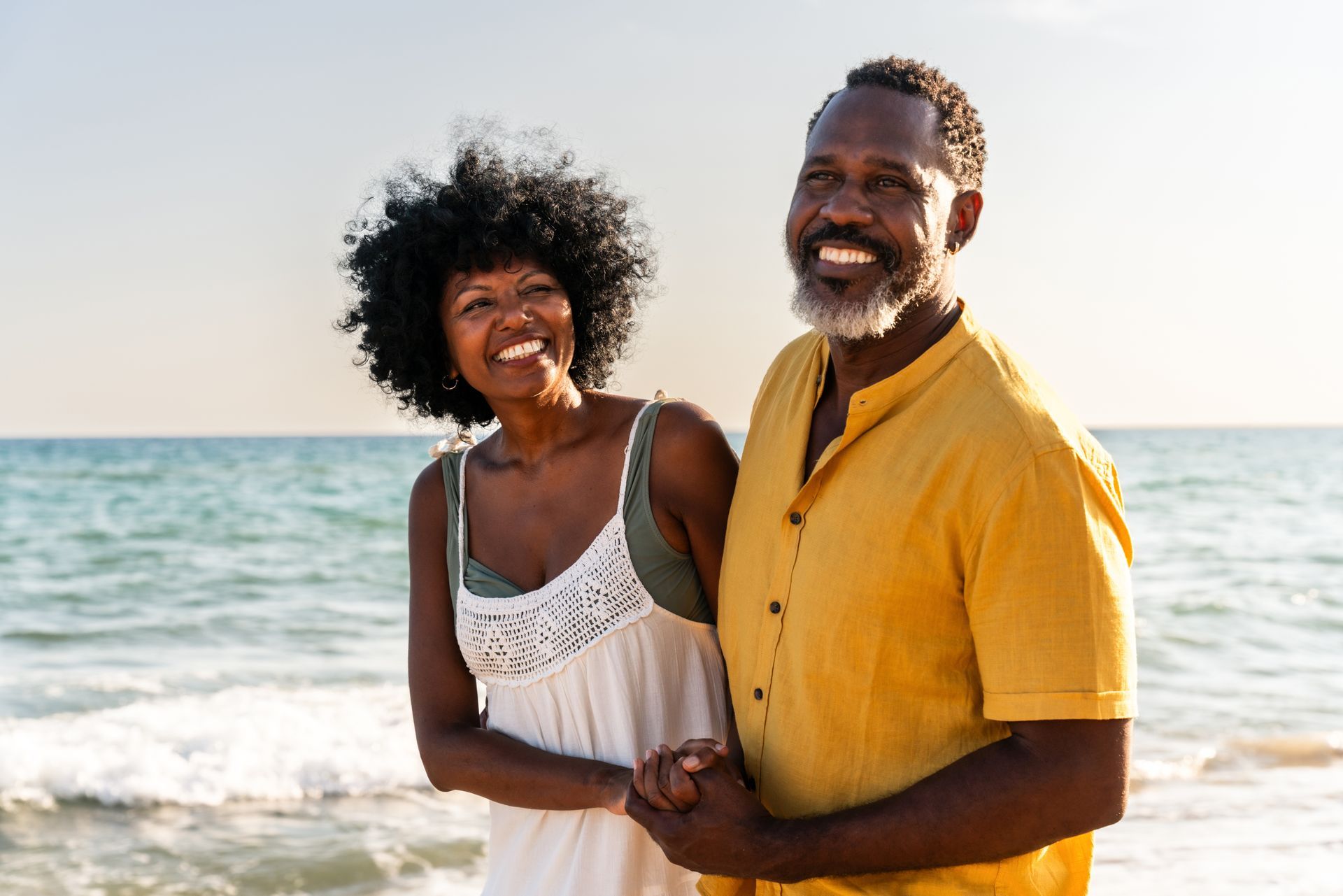 Smiling Black couple holding hands at the beach, the woman in a white dress and the man in a yellow shirt, ocean in the background.