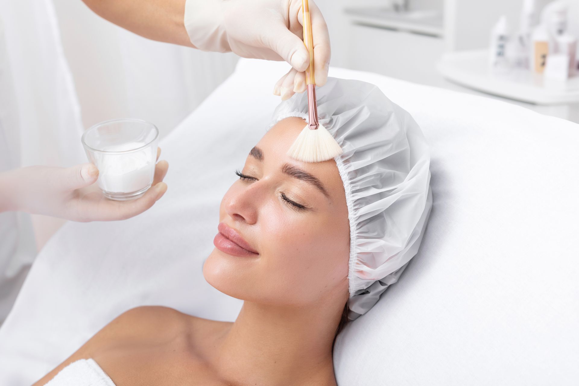 A woman receiving a facial peel at a spa. A gloved hand applies a treatment from a small bowl with a brush.