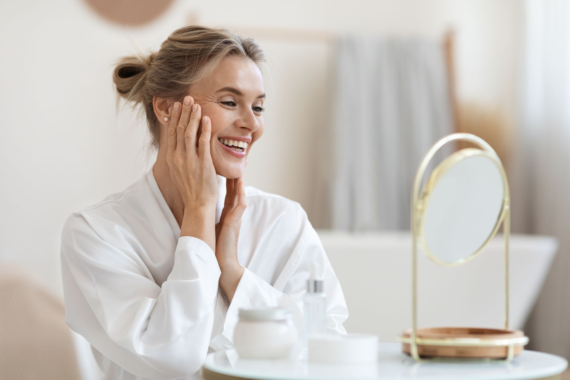 A smiling woman in a white robe examines her face in a mirror in a bright bathroom.