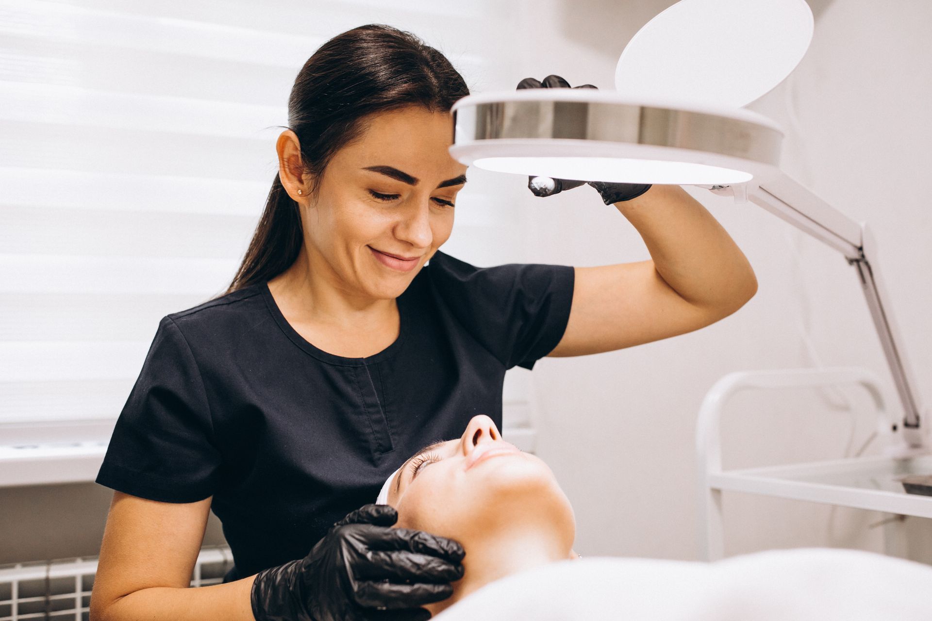 Esthetician examining a client's face under a bright magnifying lamp. Woman wears black gloves and smirks, in a bright room.