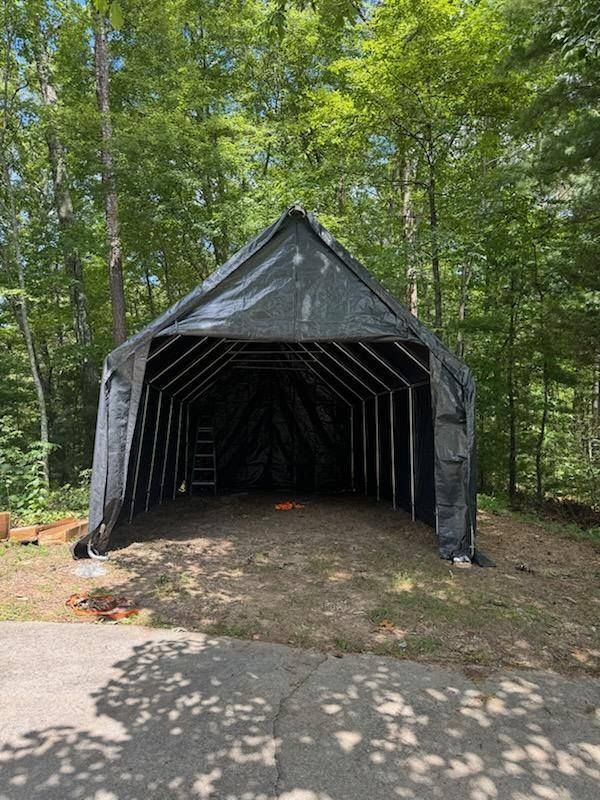 Black tarp-covered metal shelter in wooded area, open on both ends.
