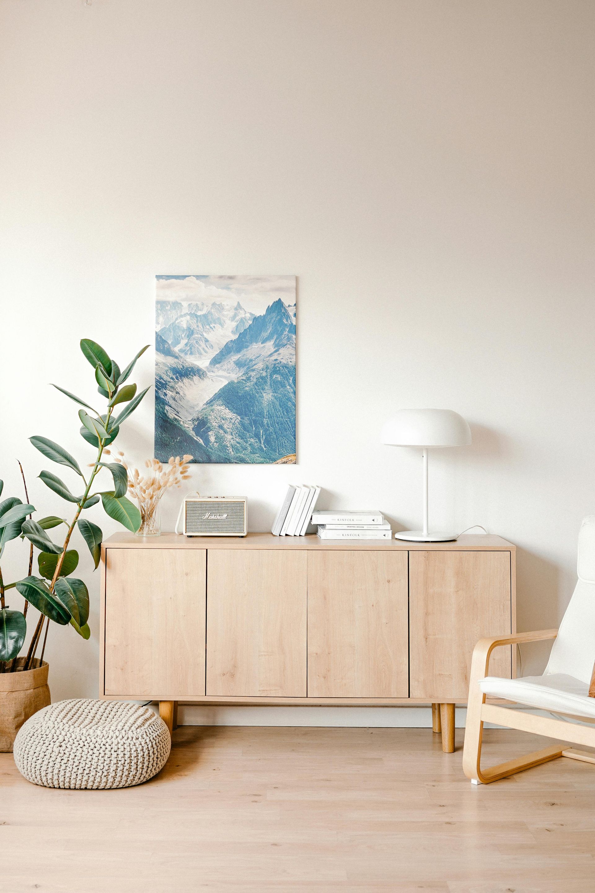 Light wood credenza with mountain art, lamp, and plants against a white wall.