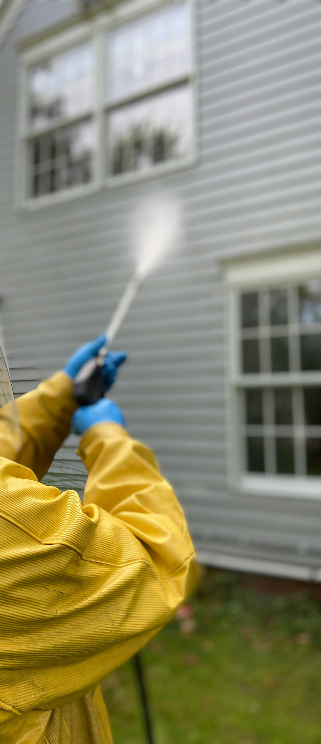 Person wearing yellow protective gear power washing a house with gray siding.