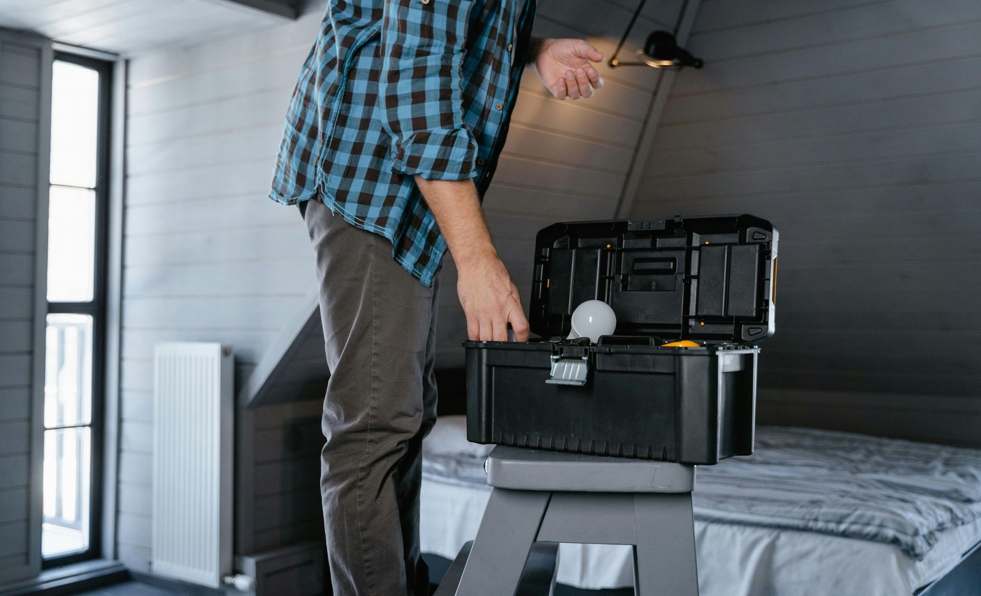 Person in plaid shirt opening a black toolbox on a stool, bedroom setting.