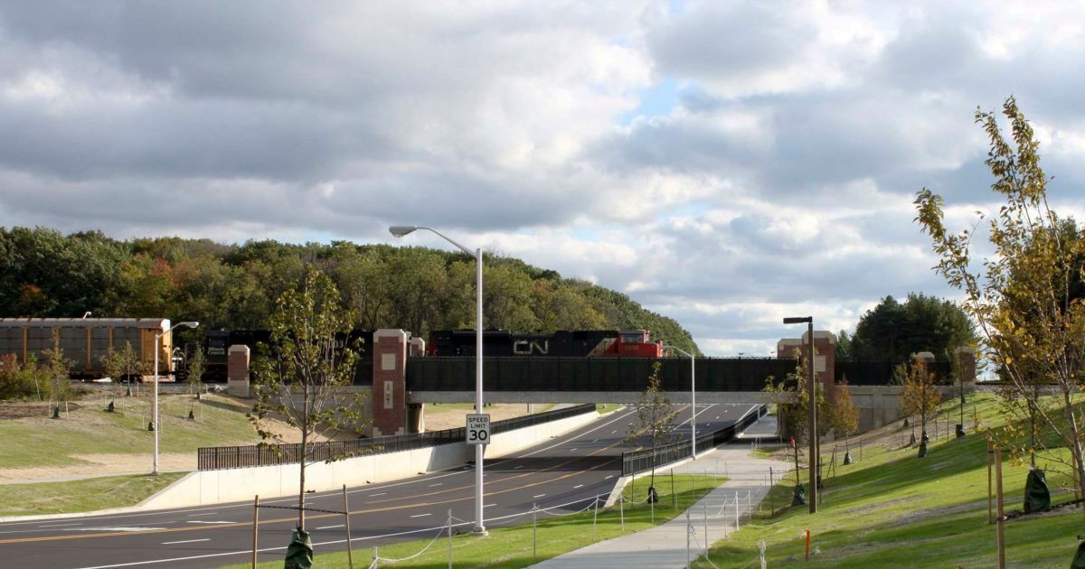 Overpass bridge over a road, with green landscape, trees and a cloudy sky.