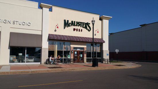 McAlister's Deli storefront with green sign. Sunny day. People at outdoor tables. Beige and brown building exterior.