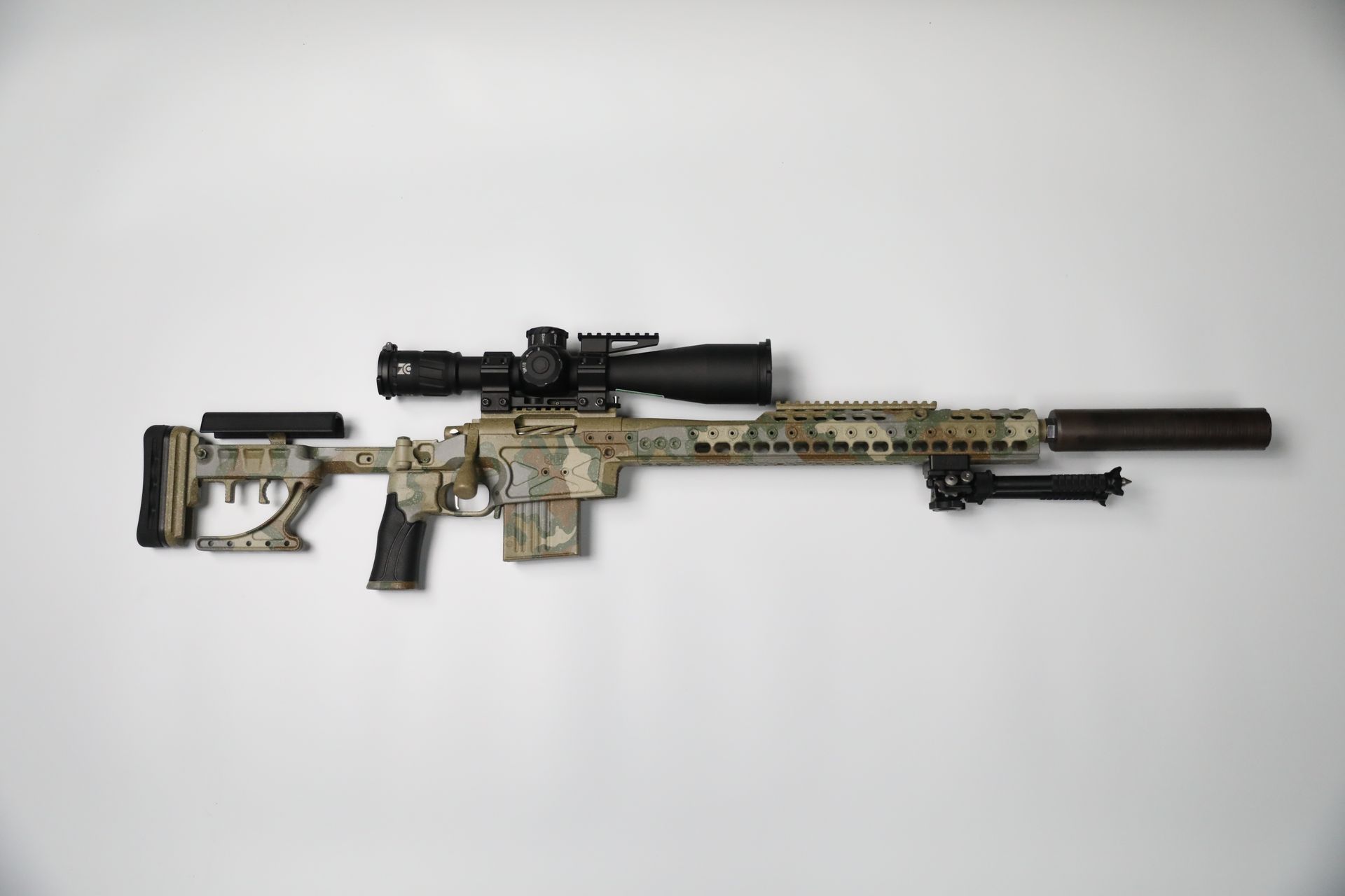 Camouflage sniper rifle with scope and suppressor against a white background.
