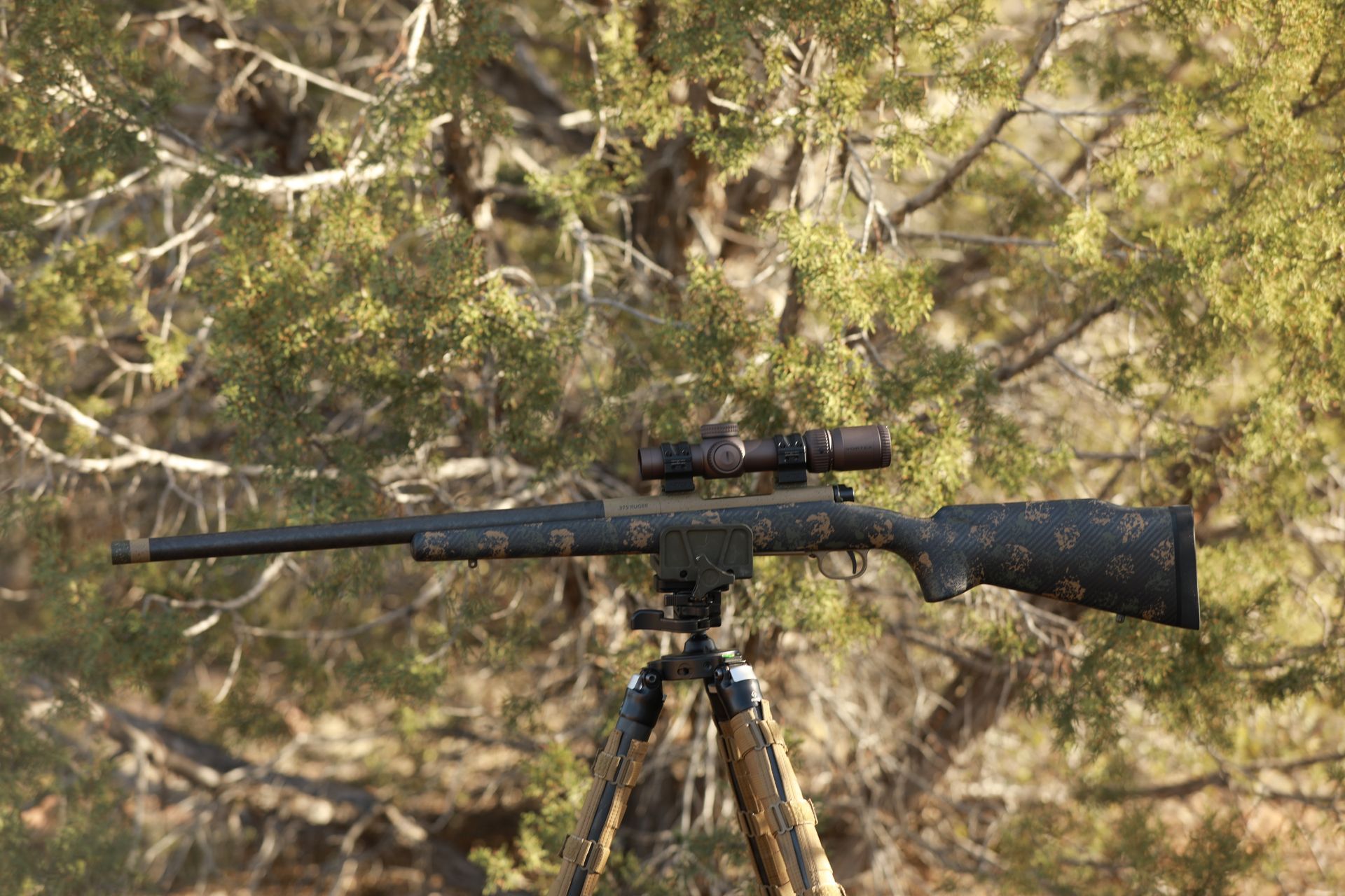 A camouflage rifle on a tripod in a wooded outdoor setting.